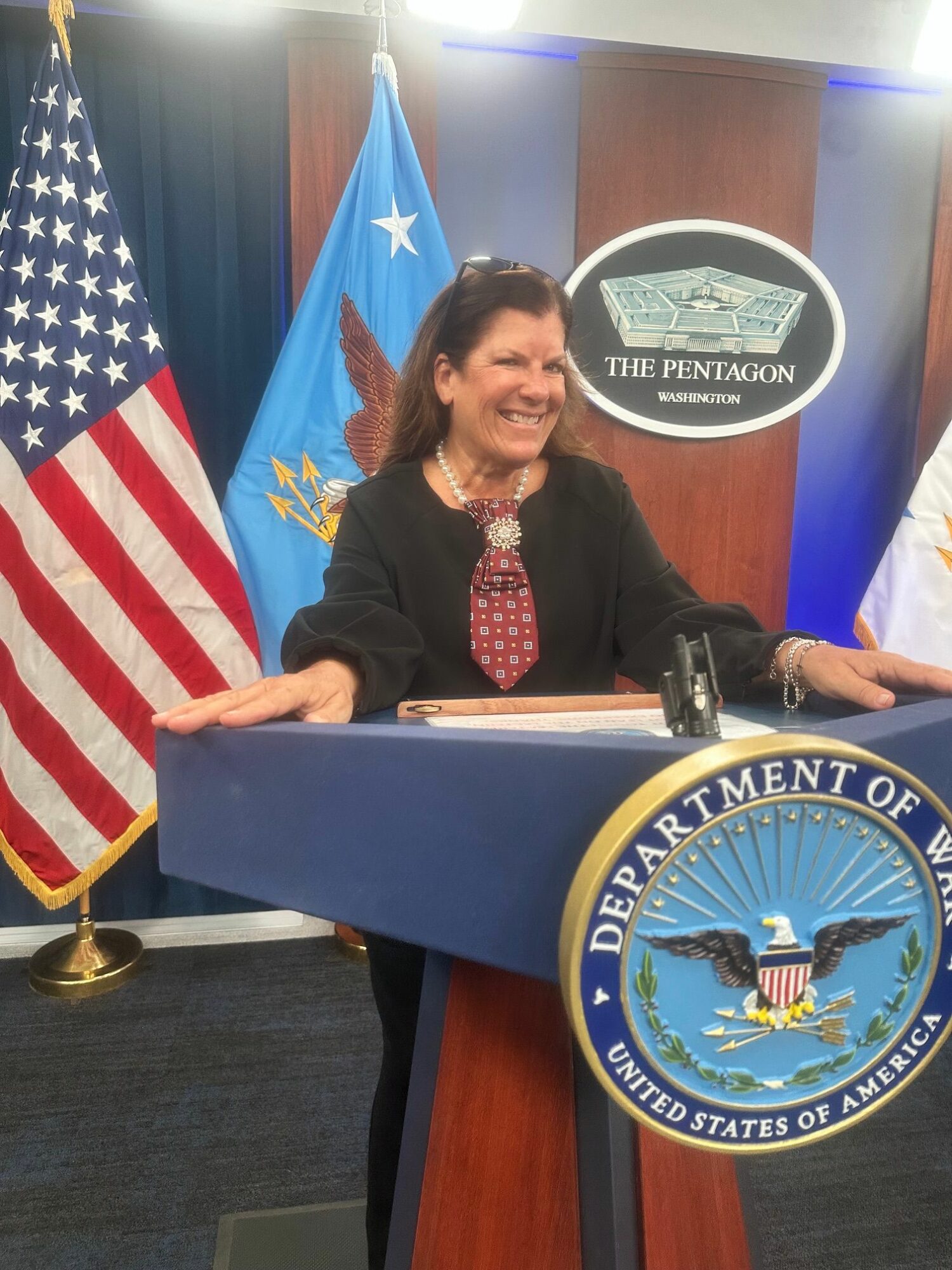 Woman speaking at a podium with the Department of War emblem, American flag, and Texas flag behind her.