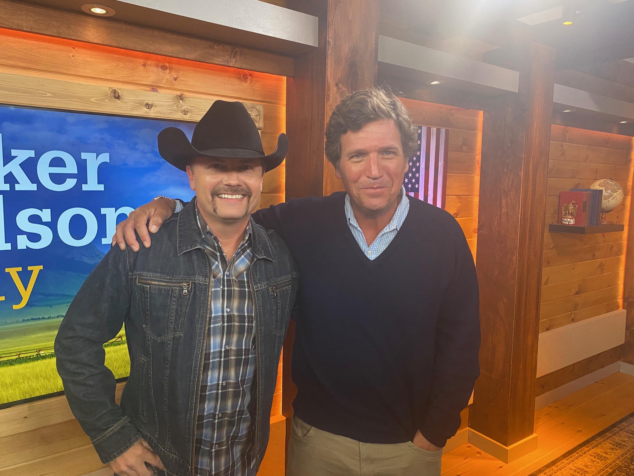 Two men stand together indoors, one wearing a cowboy hat and denim jacket, the other in a dark sweater, smiling.