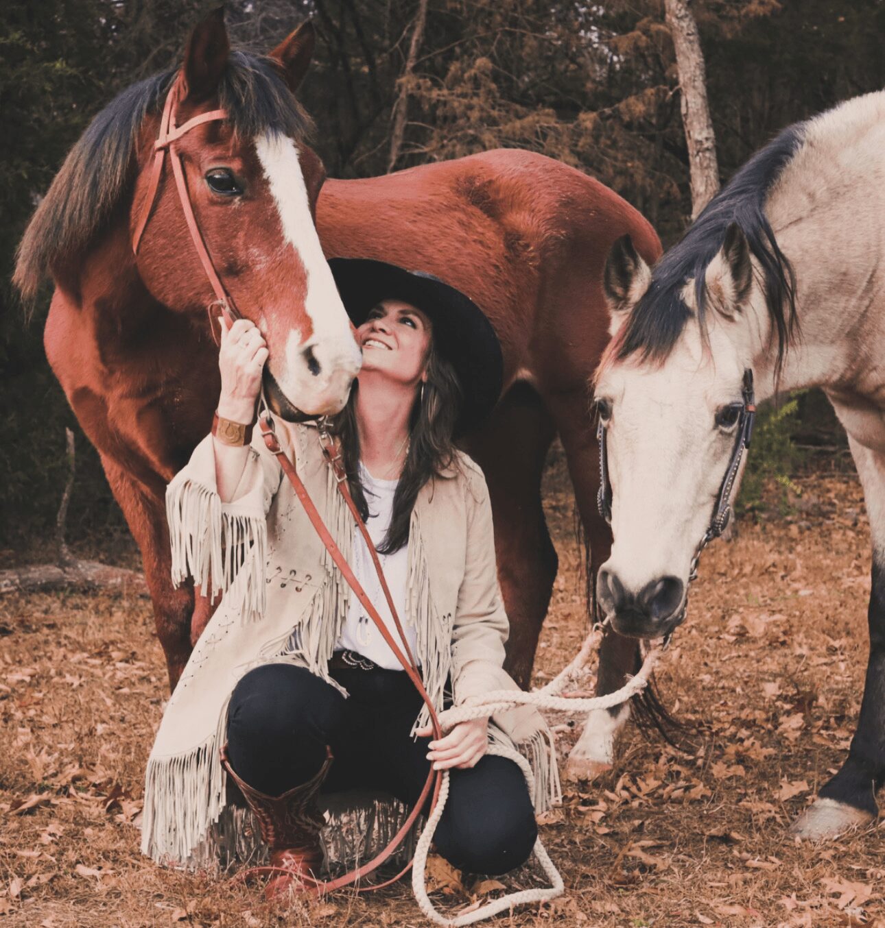 Woman sitting on ground with two horses outdoors, holding their reins, surrounded by trees and natural scenery.