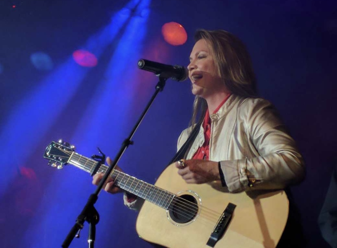 Woman singing into microphone while playing an acoustic guitar on stage with colorful lights