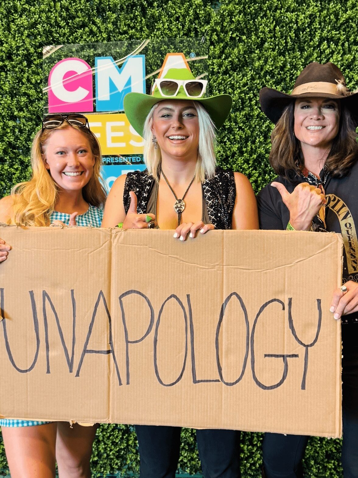 Three women holding a cardboard sign with the word 'UNAPOLOGY' written on it, smiling outdoors with a green hedge background.