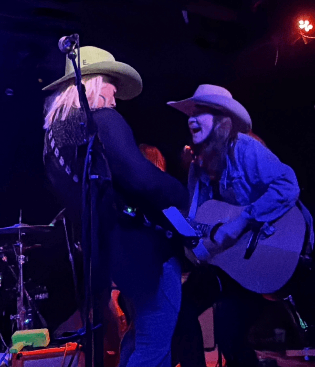 Two women wearing cowboy hats and jeans, one with a guitar, sharing a moment on stage with dark background.