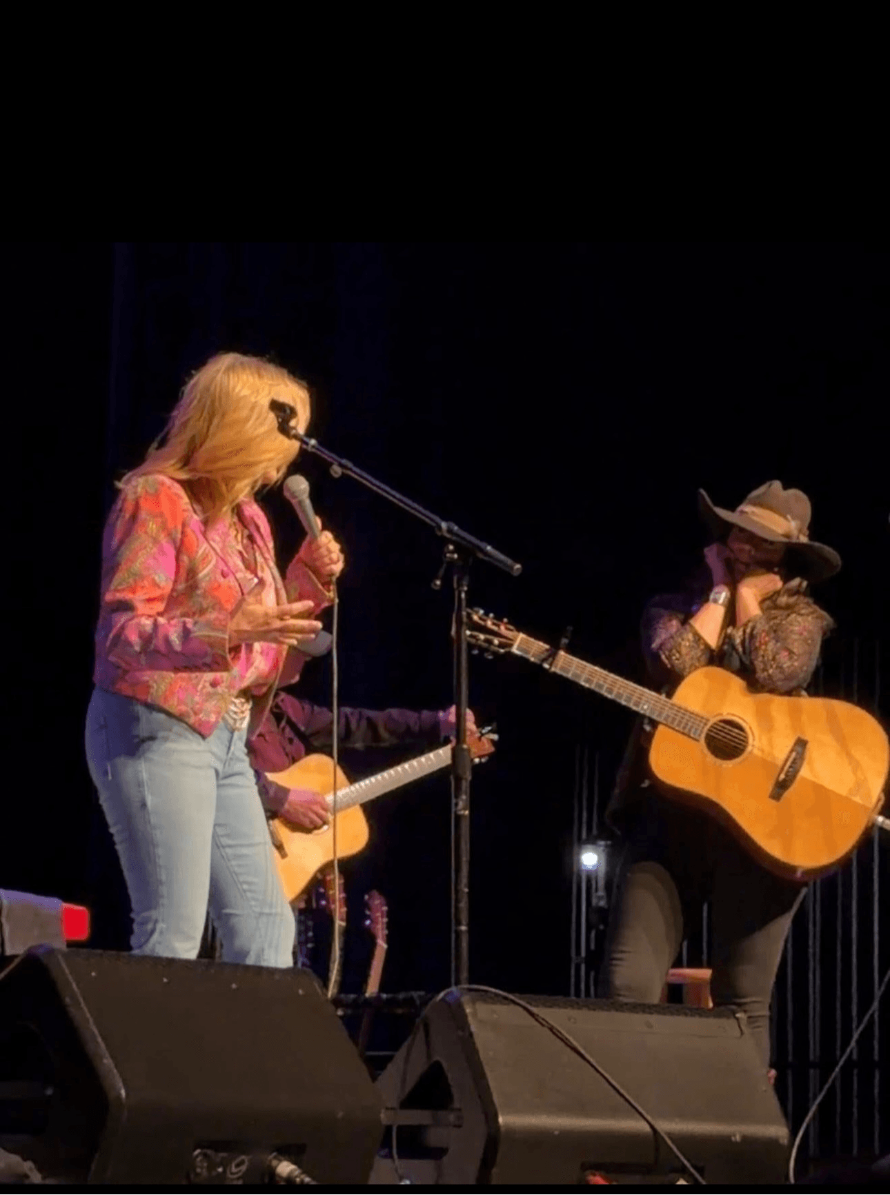Two women perform on stage with guitars, one singing into a microphone, the other playing guitar and wearing a hat.