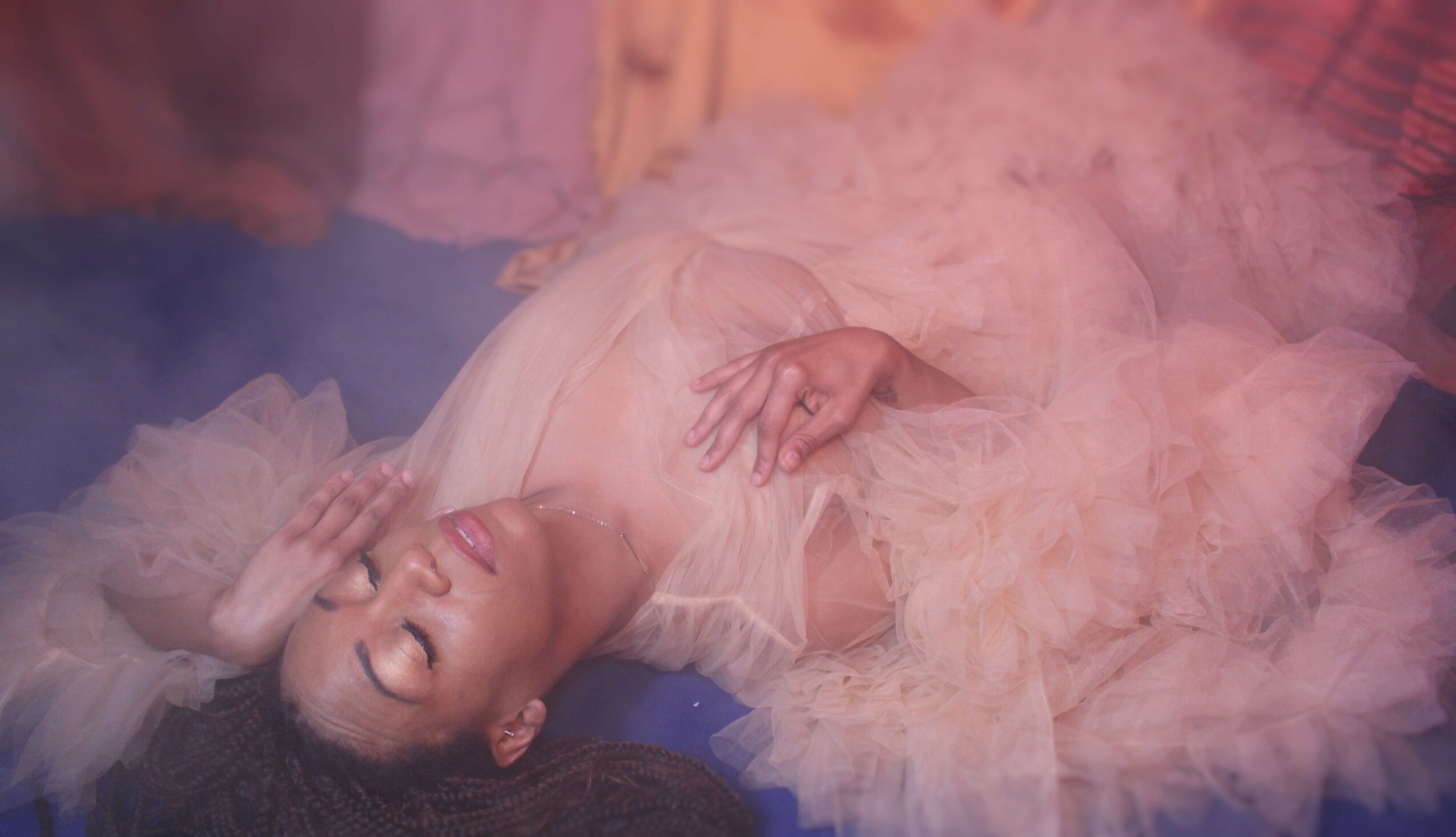 Woman lying on her back in a pink, fluffy dress, smiling with eyes closed, surrounded by pink smoke or fog.