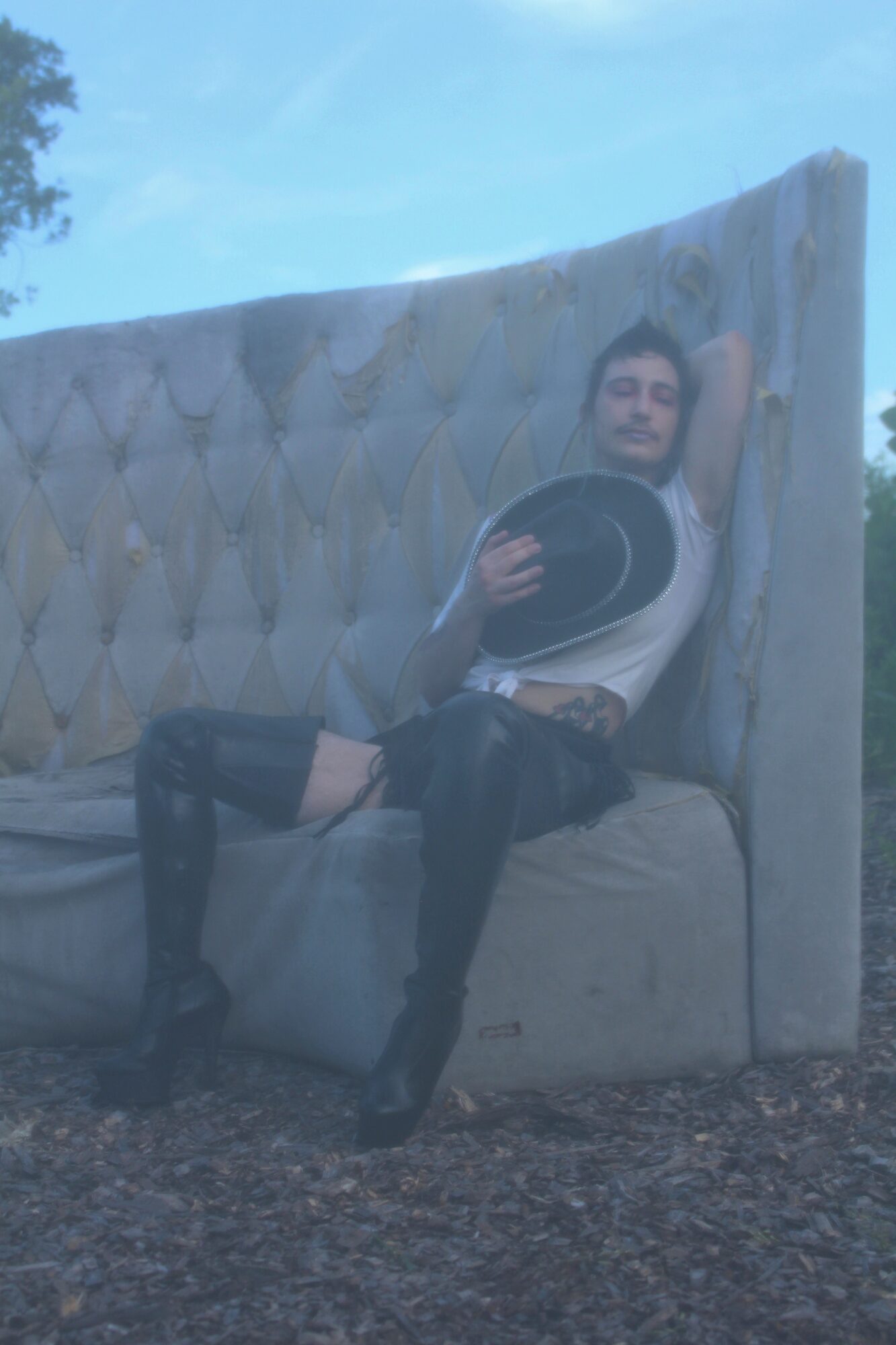 Person sitting on a large, tufted outdoor sofa holding a hat, with one arm resting on the backrest, outdoors.