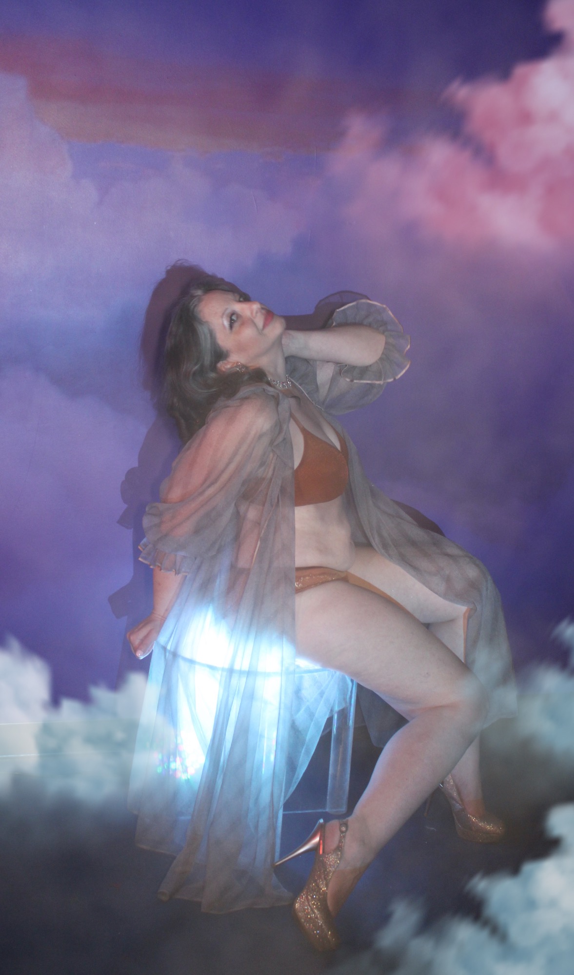 Woman sitting on a cloud-like surface with a colorful sky background, wearing a sheer robe and high heels.