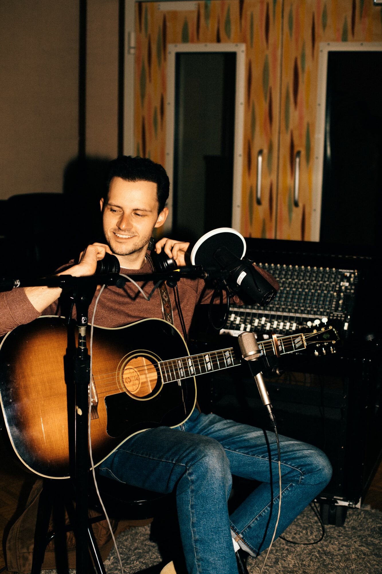 Person playing acoustic guitar in a recording studio with sound equipment and colorful wall background.