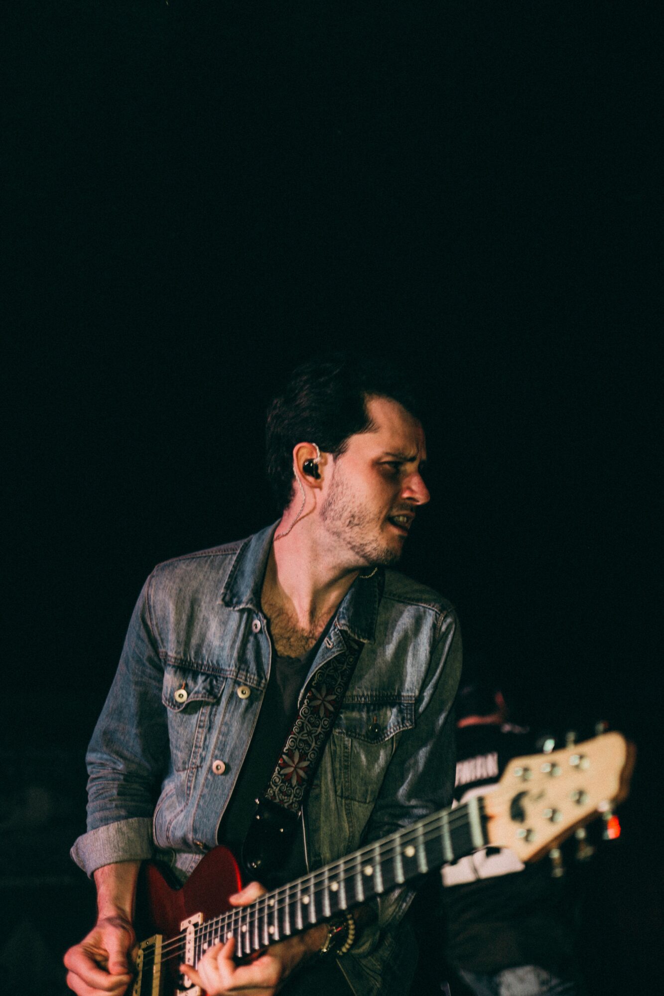 Person playing guitar on stage, wearing a denim jacket, with dark background.