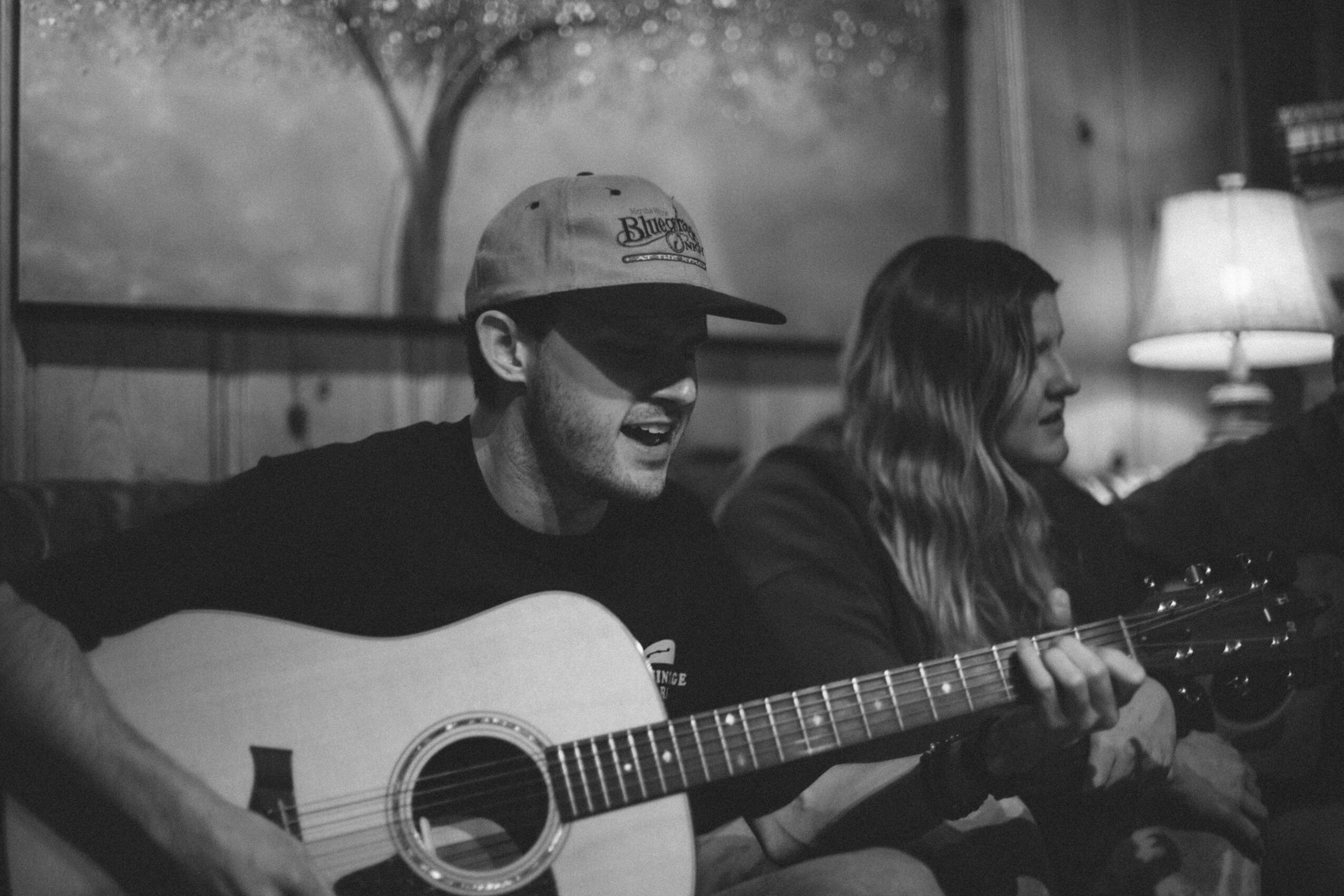 Two people sitting indoors, one playing guitar, with a lamp and window in the background.