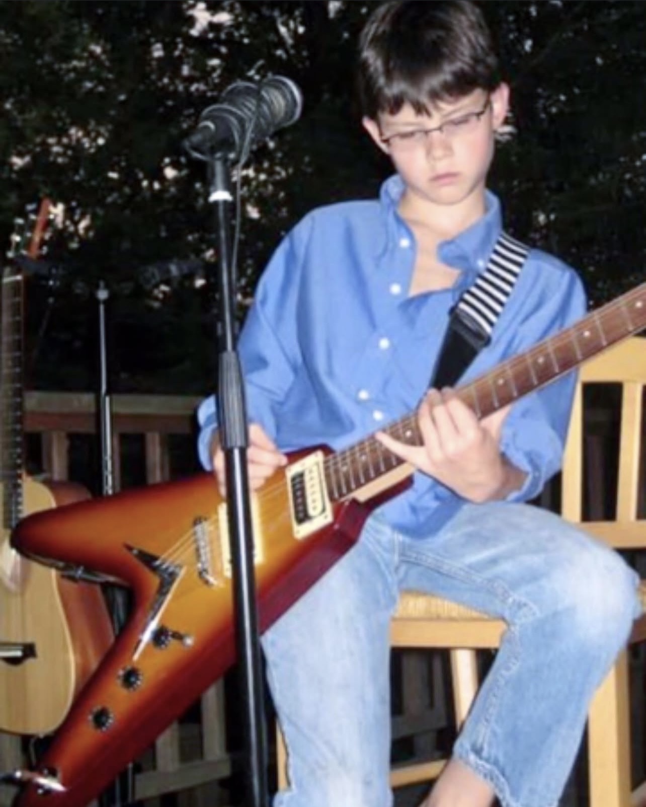 Young boy with glasses playing an electric guitar outdoors, sitting on a wooden chair, with a microphone nearby.