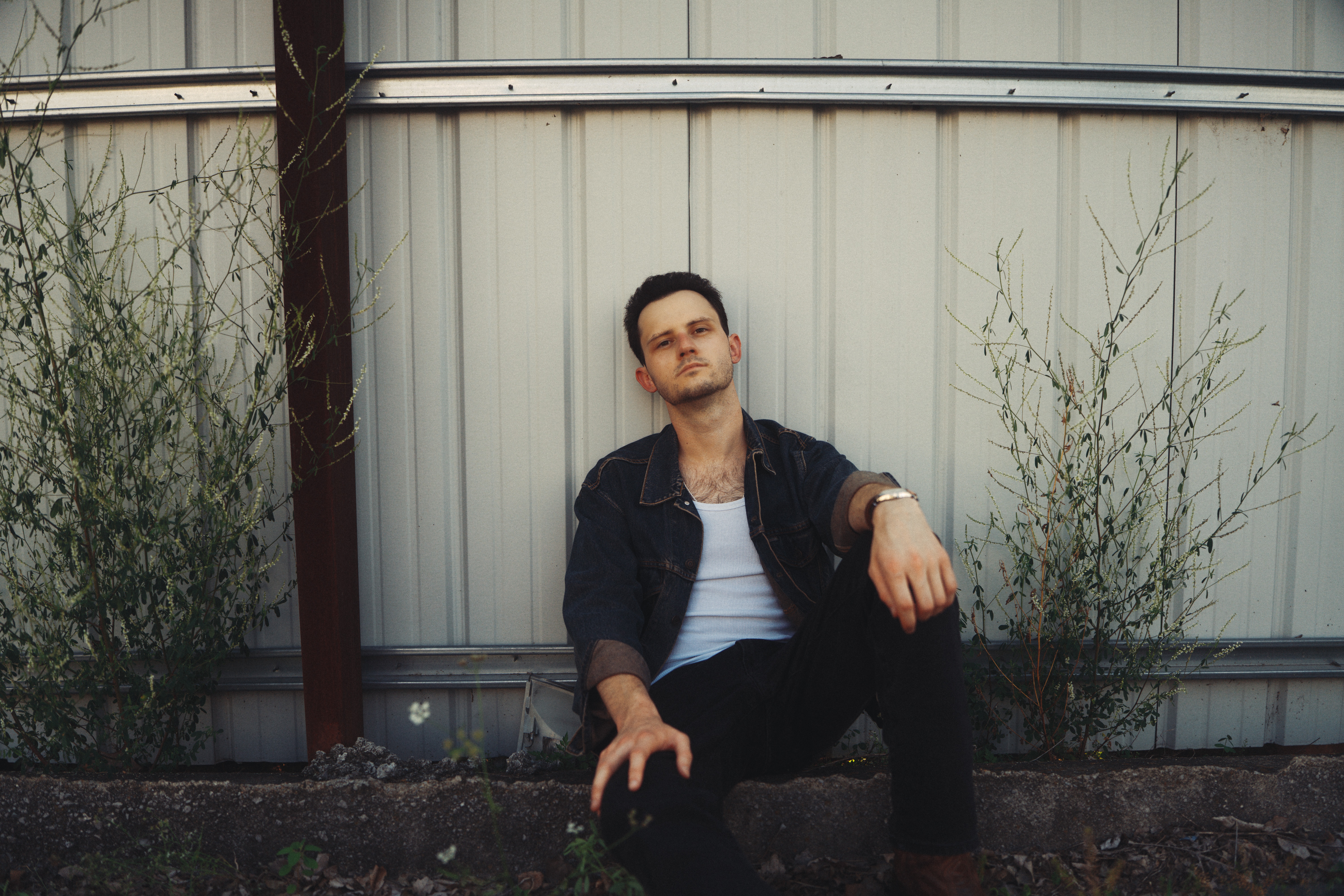 Young man sitting outdoors in front of a metal wall with plants on either side.
