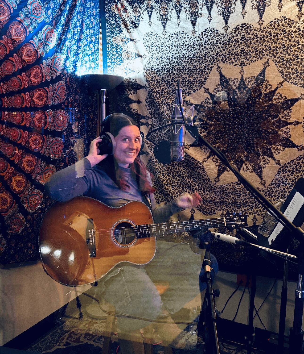 Woman with headphones playing guitar in a room with colorful tapestry backgrounds, microphone, and music stand.