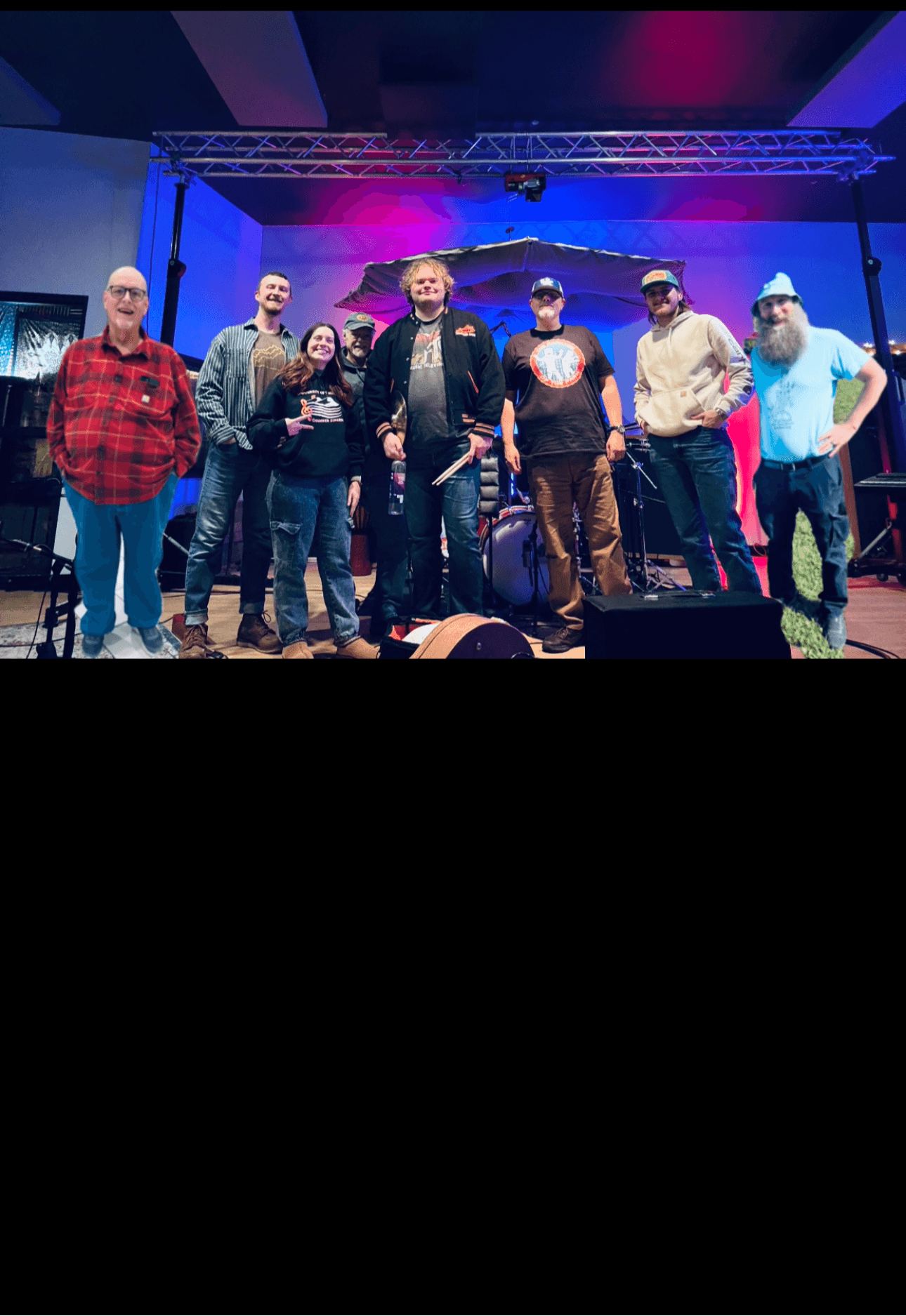 Group of nine people standing on stage with colorful lighting and equipment in background.