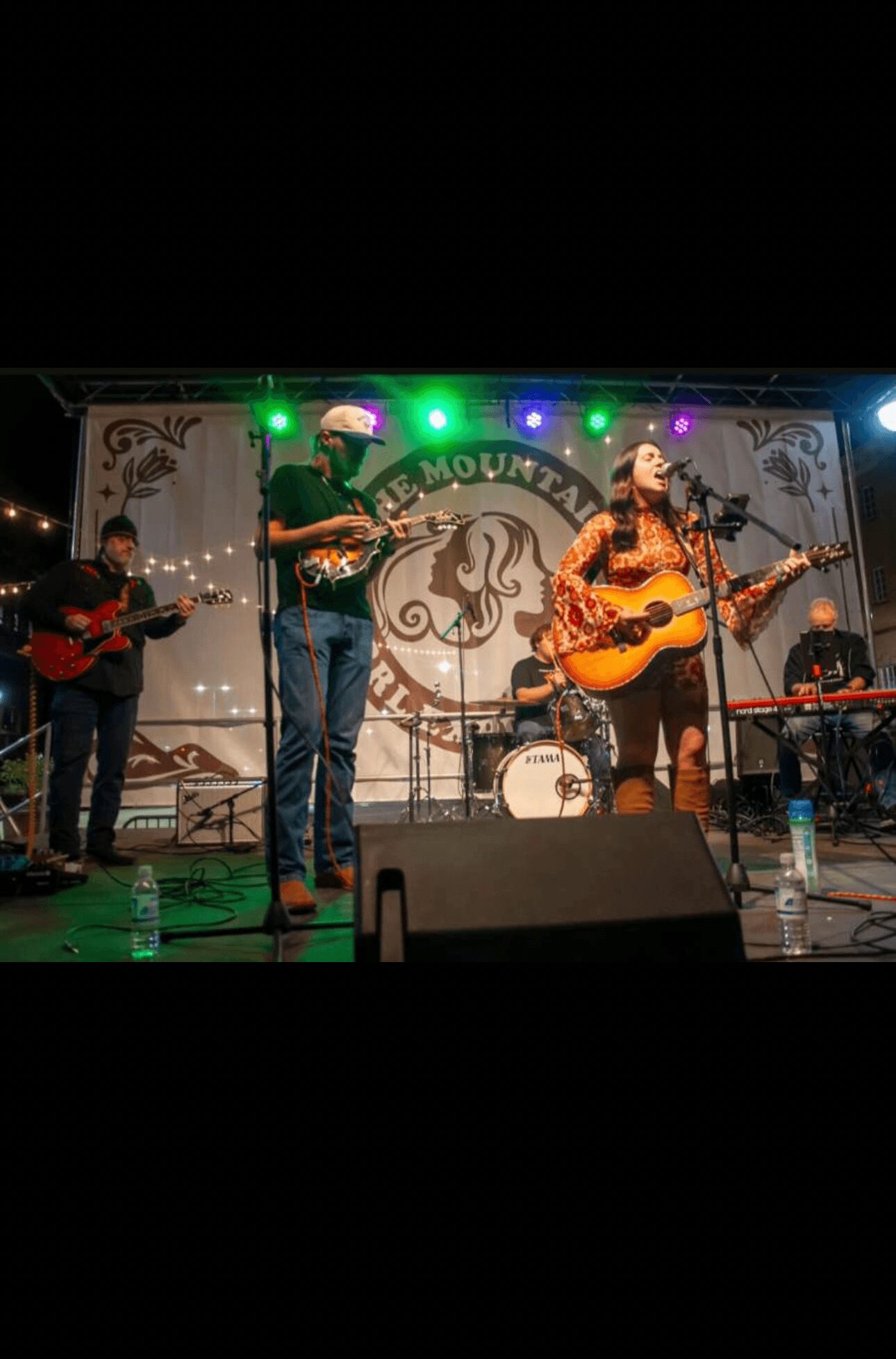 Band performs on stage with four musicians playing guitars and singing, colorful stage lights, and a backdrop with a logo.