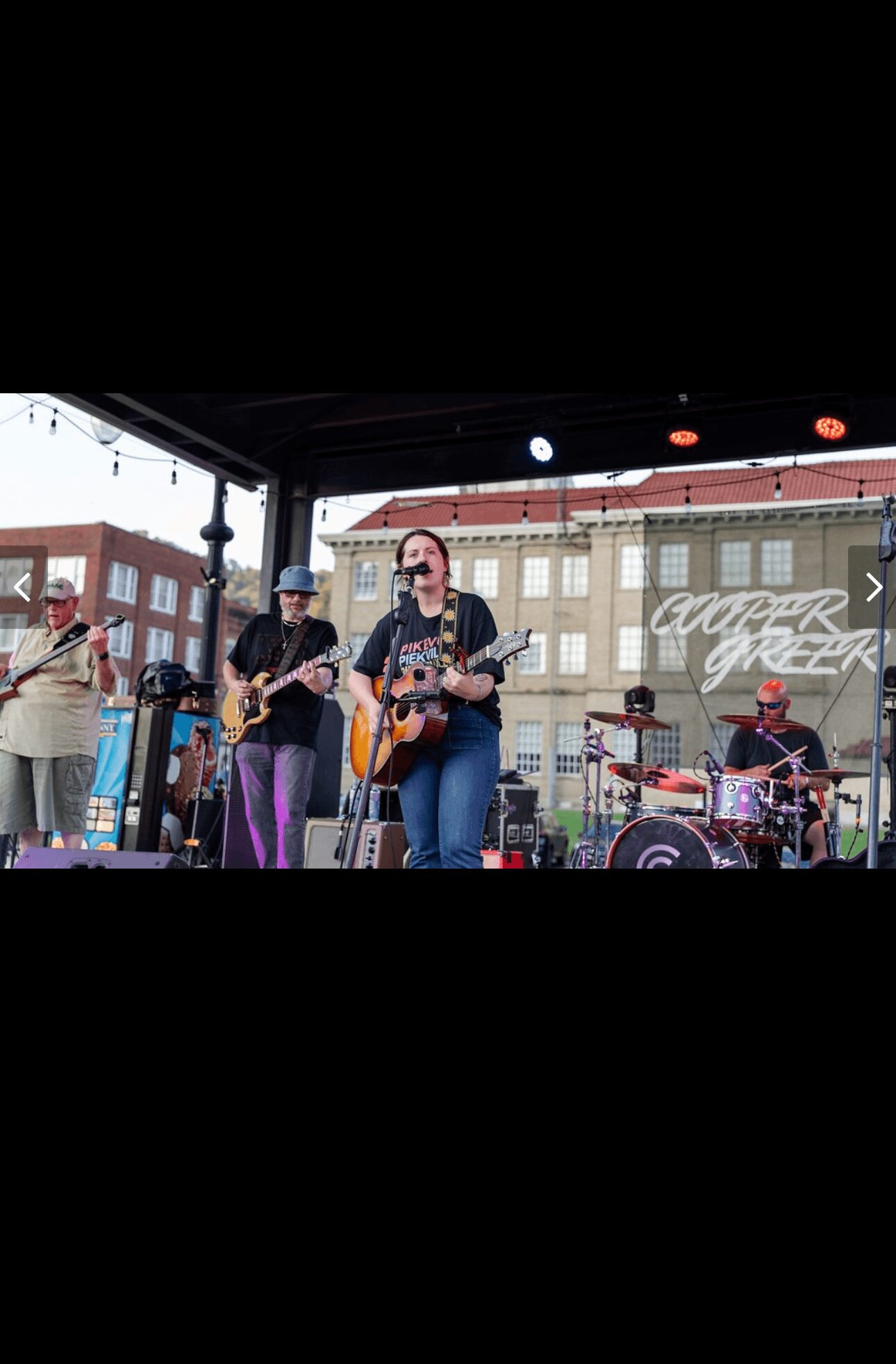 Band performs on stage outdoors with buildings in background, musicians playing instruments, some wearing hats, audience visible.