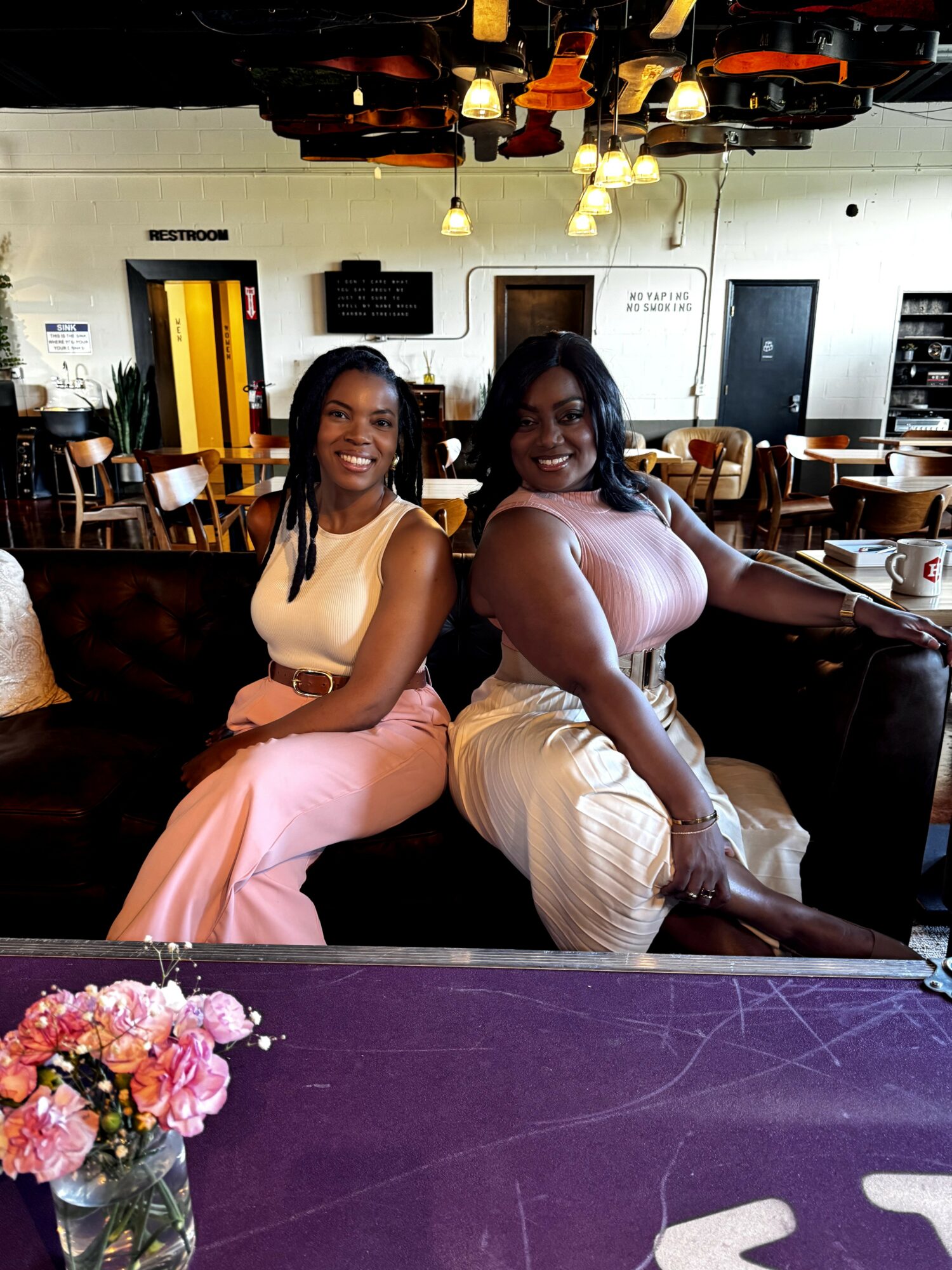 Two women sitting on a couch in a cafe, smiling, with a purple table and flowers in the foreground.
