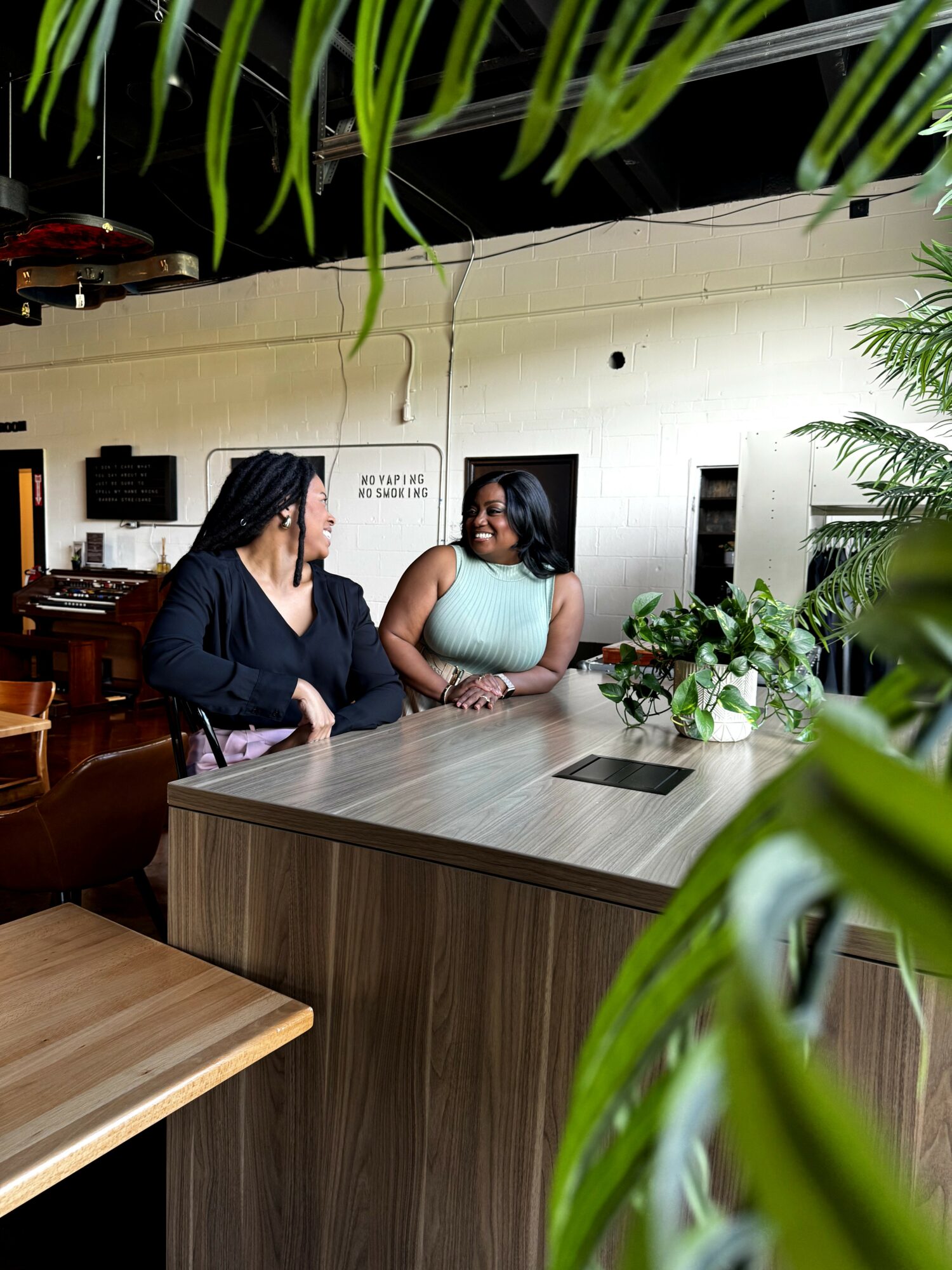 Two women sit at a table in a room with plants and a white wall, engaged in conversation.