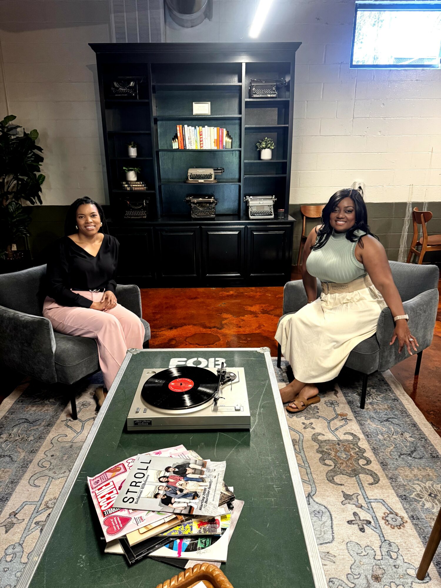 Two women sit on armchairs in a room with a black bookshelf, a table with magazines and a record player, and a patterned rug.