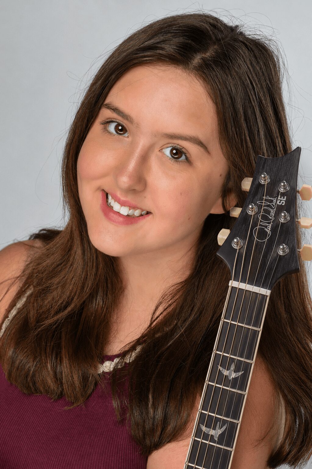 Young woman with long brown hair smiling, holding a guitar headstock, wearing a maroon top, against a light background.