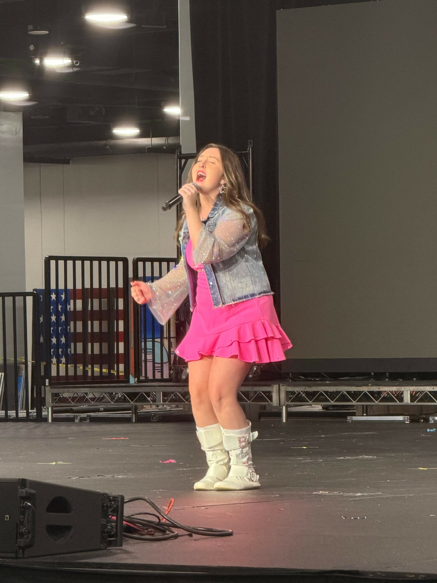Young girl with long hair in a pink skirt, gray top, and white boots standing on stage, holding a microphone.