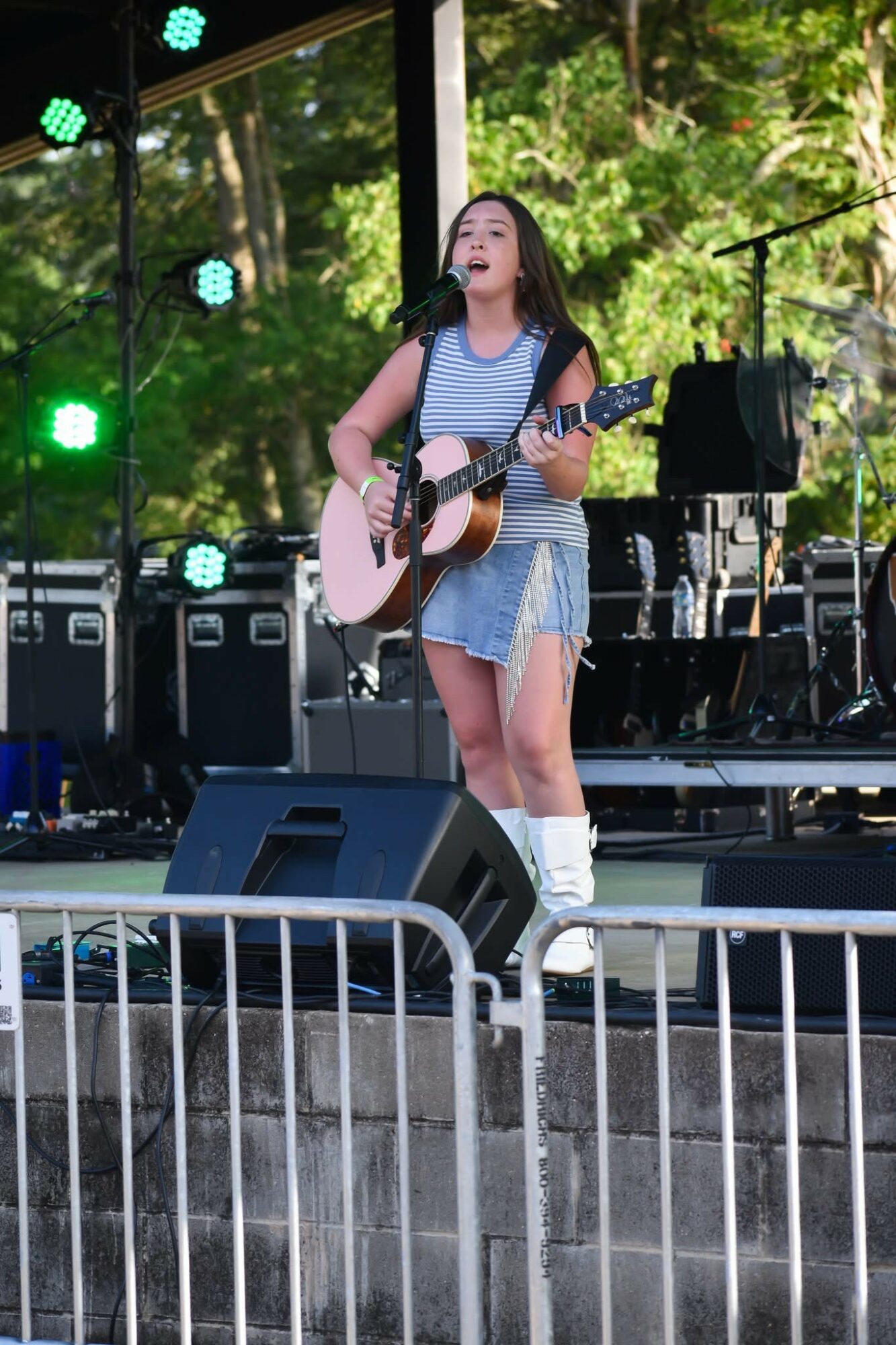 Young woman singing and playing guitar on outdoor stage with trees in background, stage equipment visible.