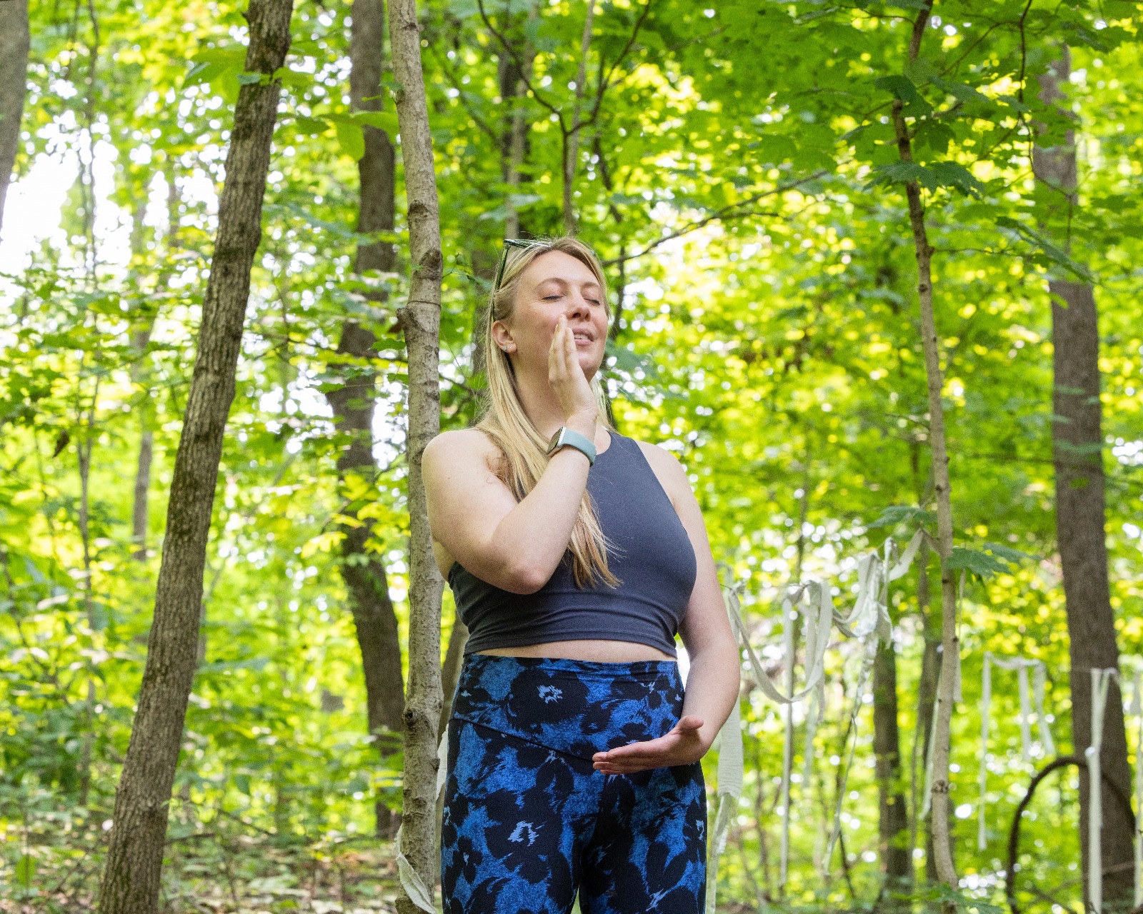 Young woman with long blonde hair standing in a green forest, eyes closed, hand near face, wearing a black crop top and patterned pants.
