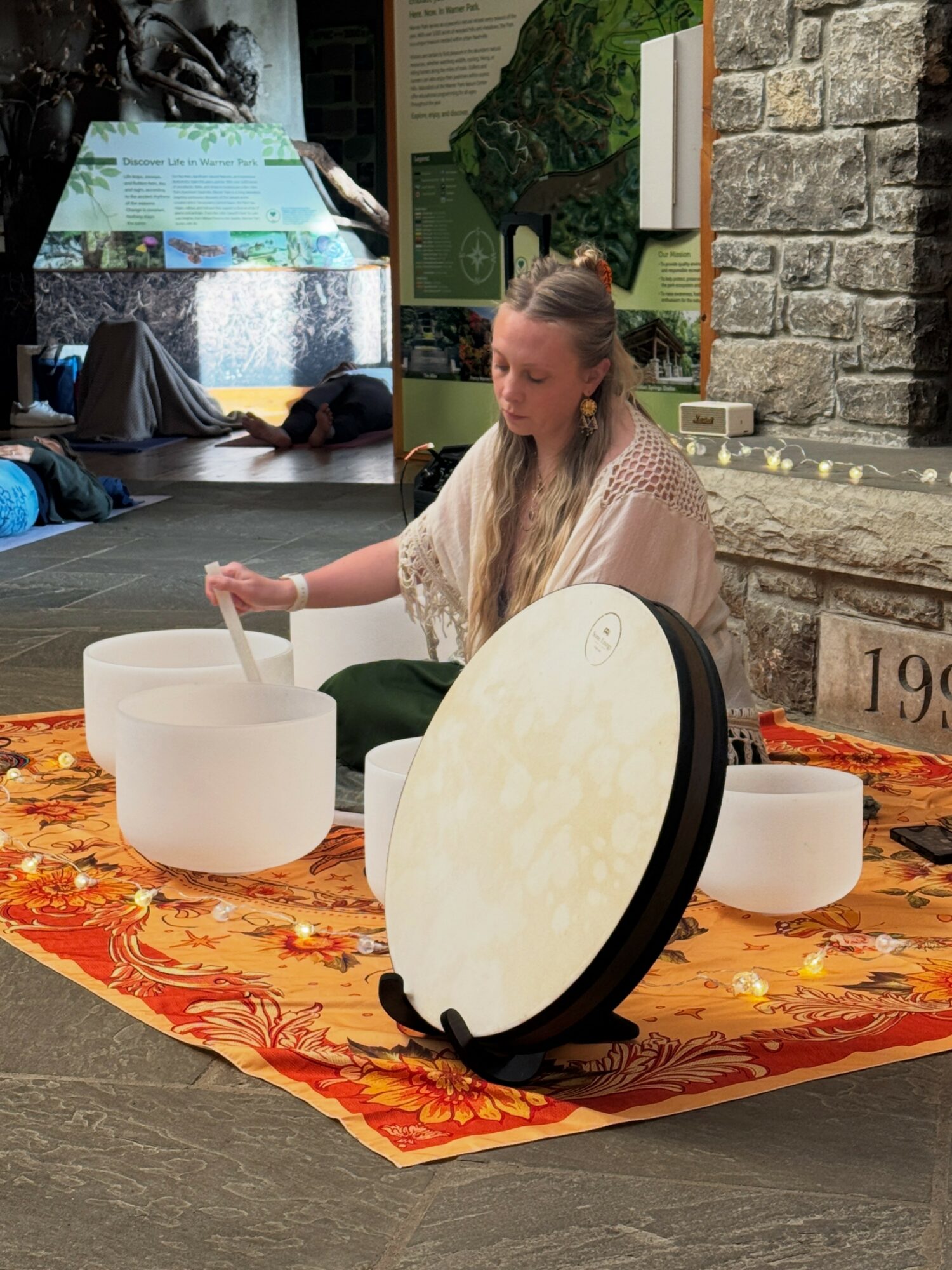Person playing singing bowl with other bowls on a colorful cloth, indoor setting with display boards and brick wall