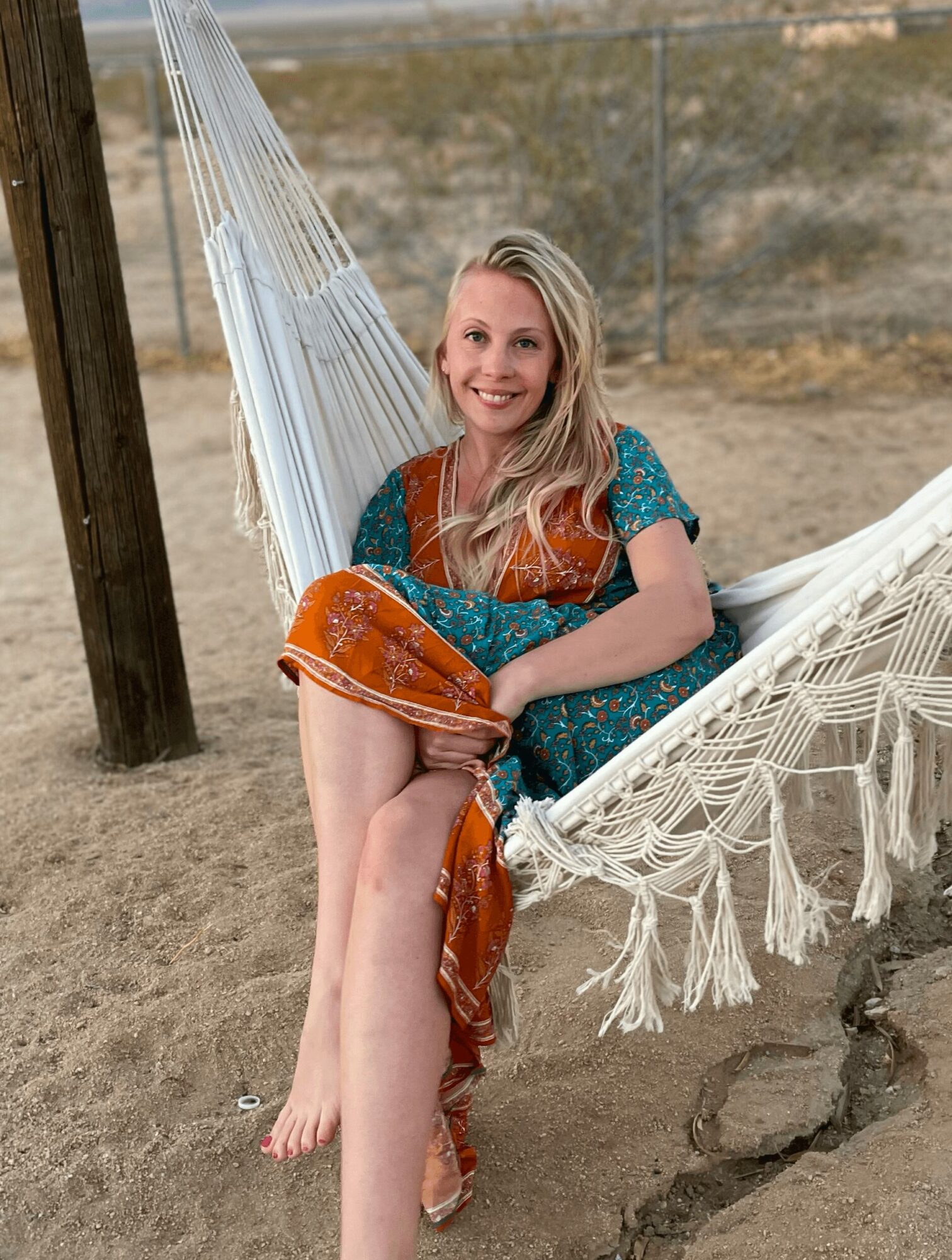 Woman sitting on a hammock at the beach, smiling, holding a bag, with sand and a wooden post nearby.
