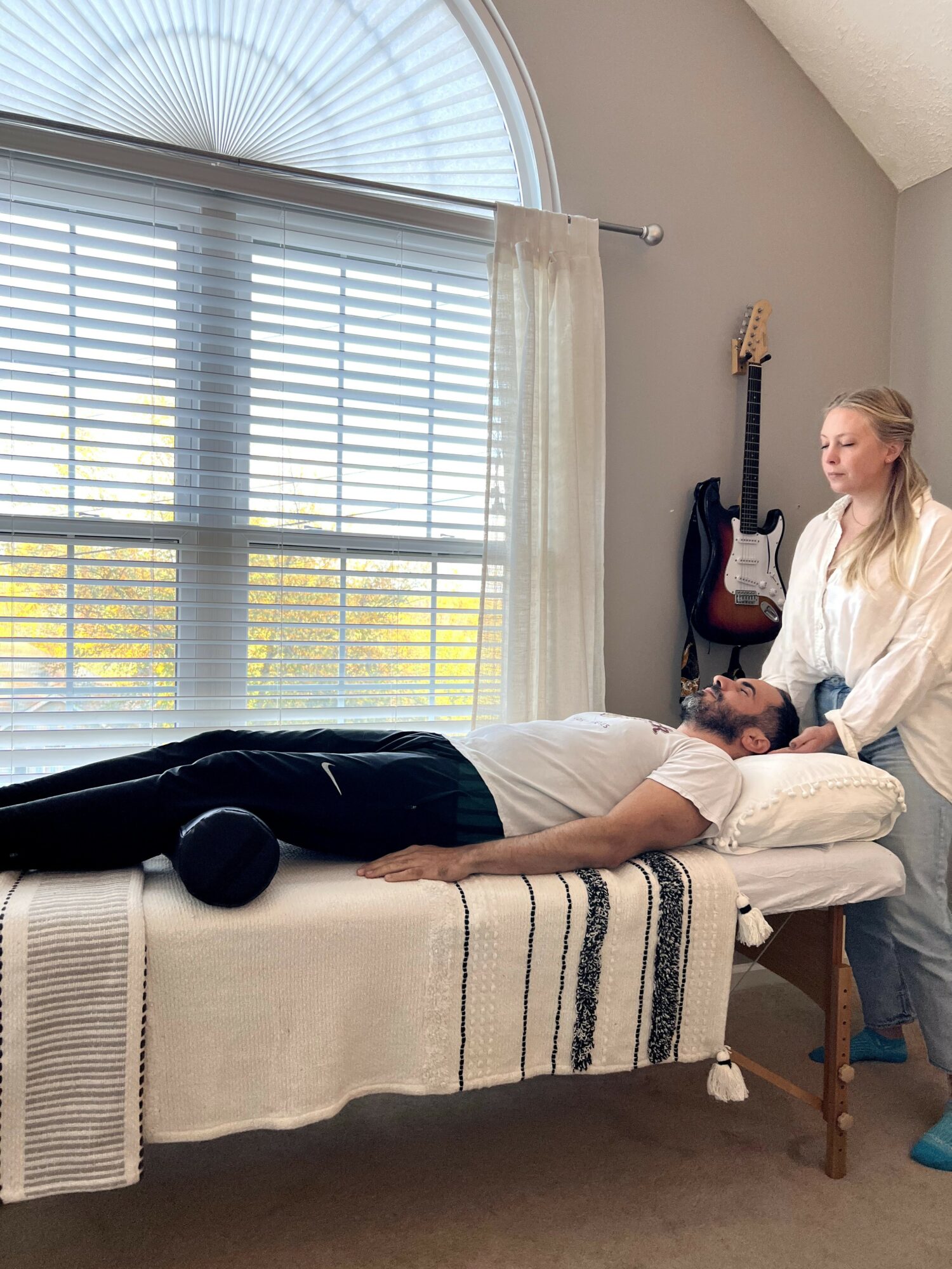 Person lying on a bed with a woman standing beside, in a room with large window and guitars on the wall.