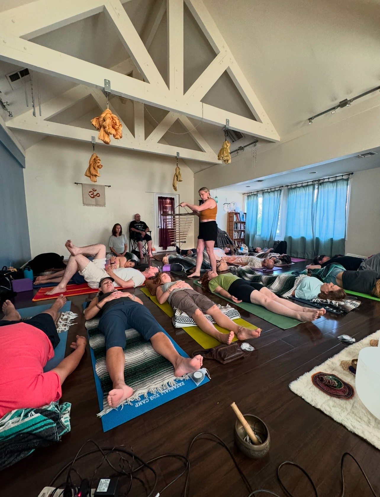 Group of people lying on mats in a room with a person standing and leading a session, blue curtains, and ceiling beams.
