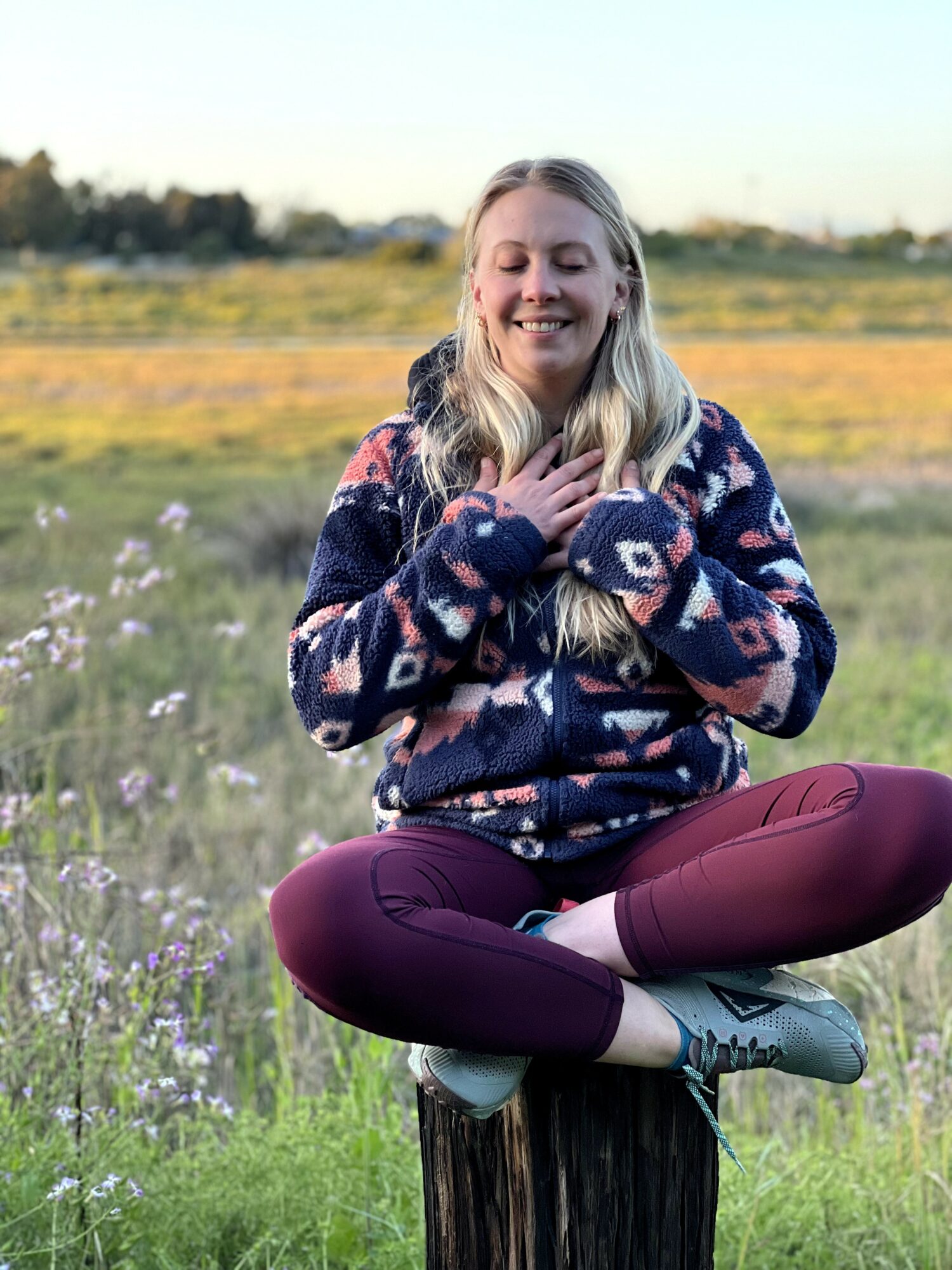 Woman sitting cross-legged on a tree stump in a field, smiling with eyes closed, wearing a patterned sweater and maroon pants.