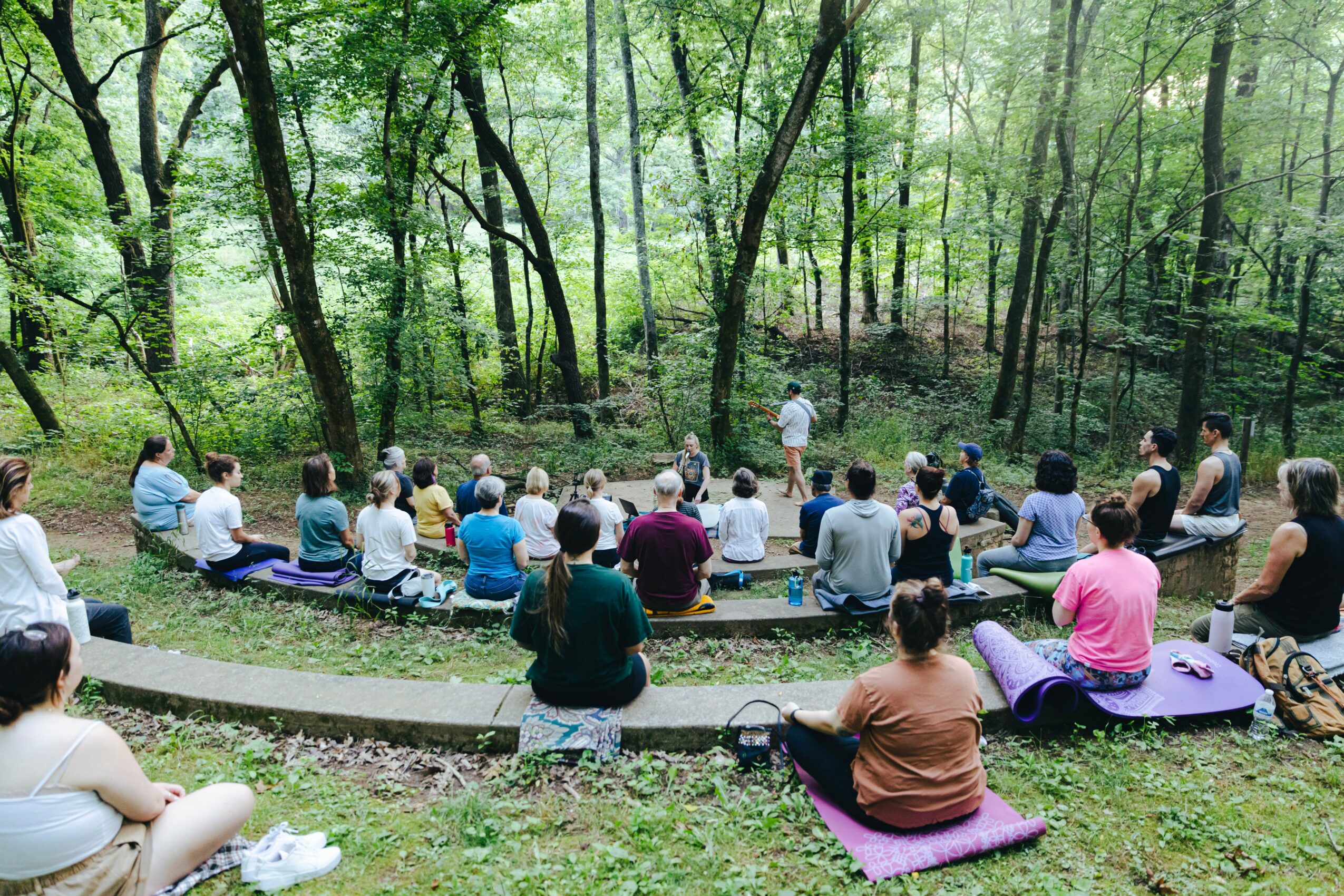 Group of people sitting on purple mats outdoors in a forest, listening to a person speaking in front of trees.