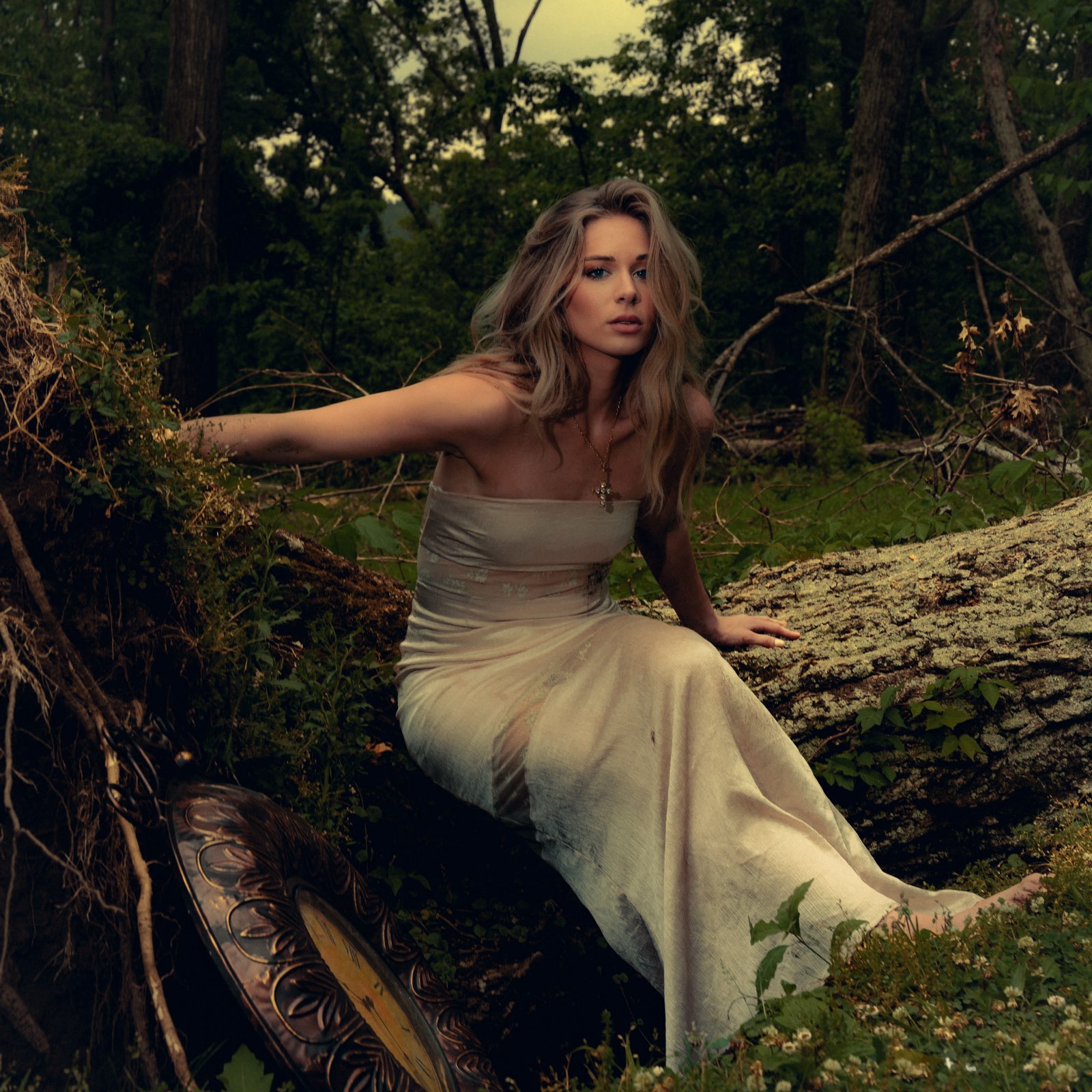 Woman in a white dress sitting outdoors on a fallen log in a forested area.