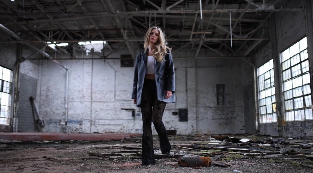 Woman standing in an abandoned industrial space with large windows and debris on the floor.