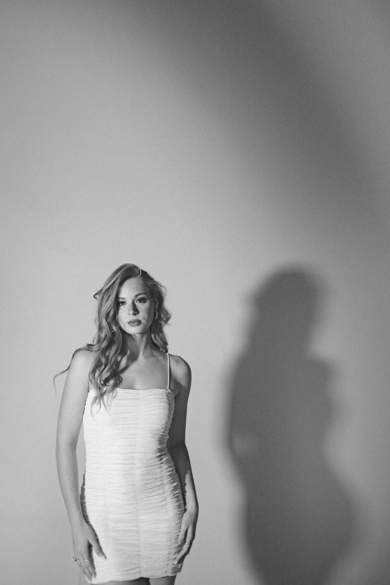 A woman in a fitted dress stands against a plain background with a shadow cast on the wall.