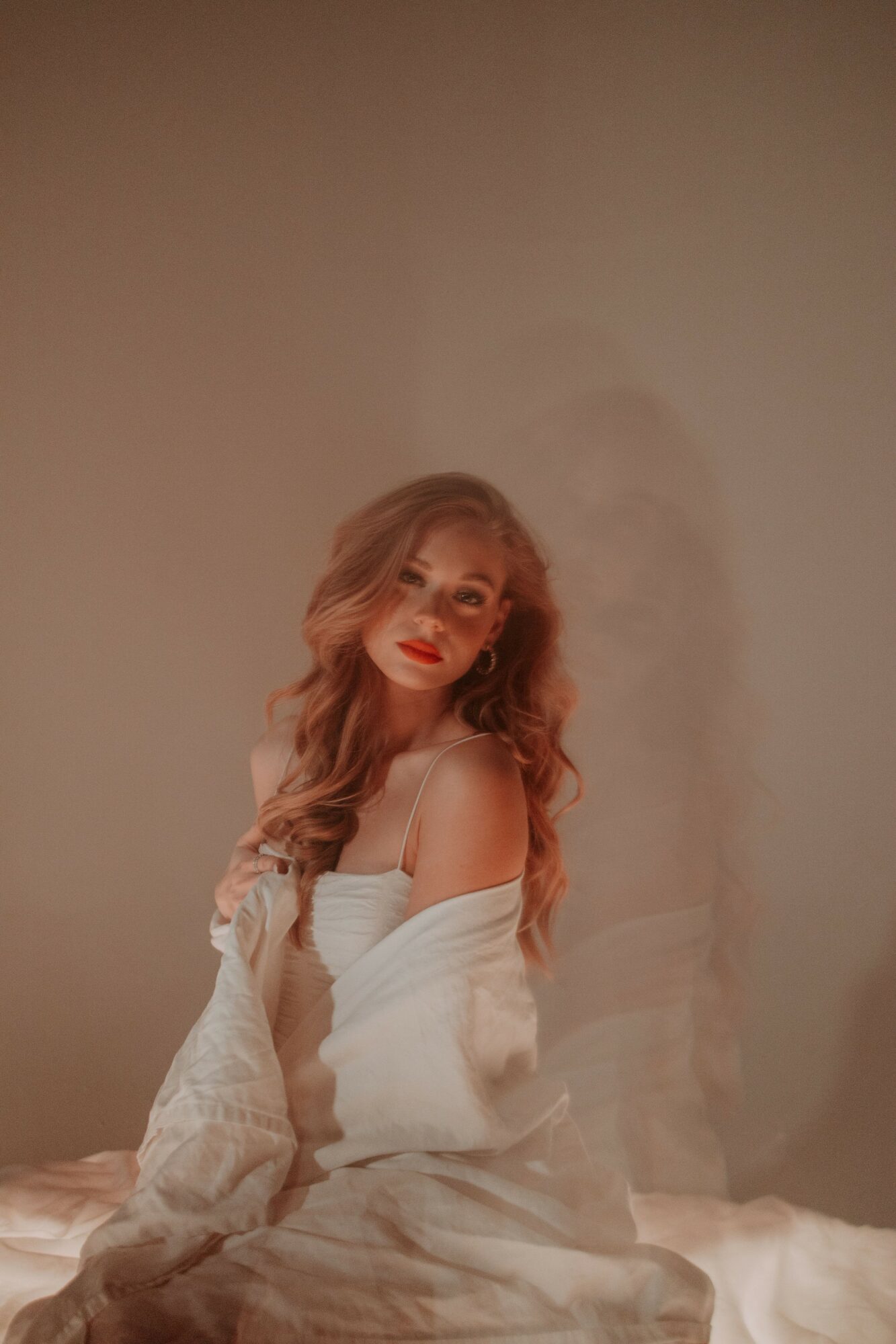 Young woman with long, wavy hair and red lipstick, wearing a white top and a light-colored cardigan, sitting on a bed.