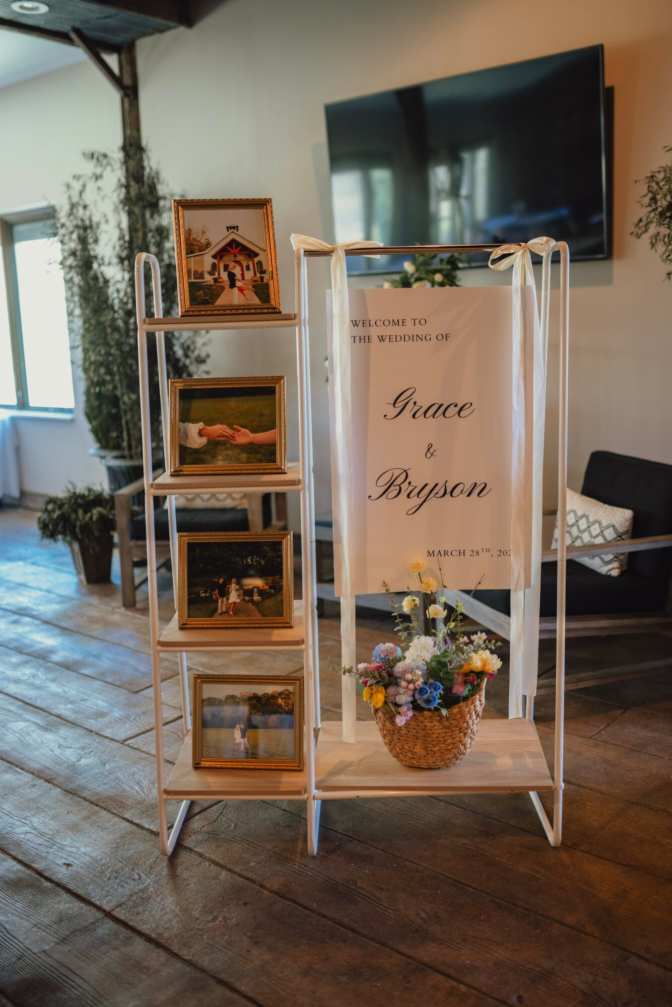 Display stand with framed photos, a welcome sign, and a flower basket in a room with a TV and window.