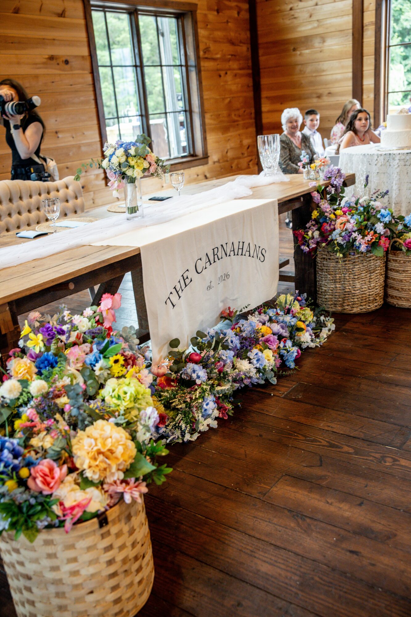 Wedding reception with floral arrangements, wooden walls, and a table with a sign reading 'The Carvahars,' guests seated in the background.