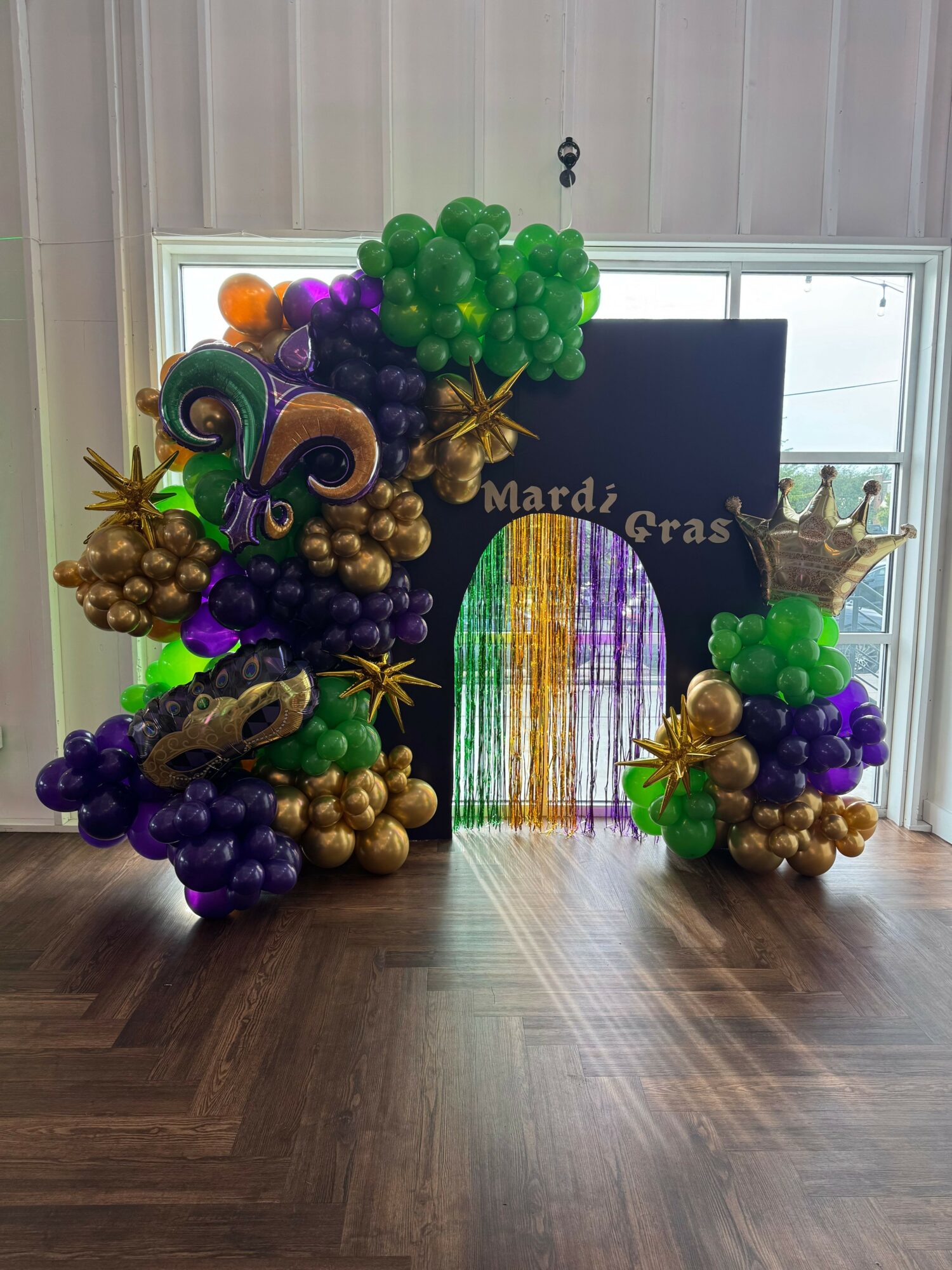 Colorful balloon arrangement with purple, green, and gold balloons, a black backdrop, and a Mardi Gras sign, on a wooden floor.