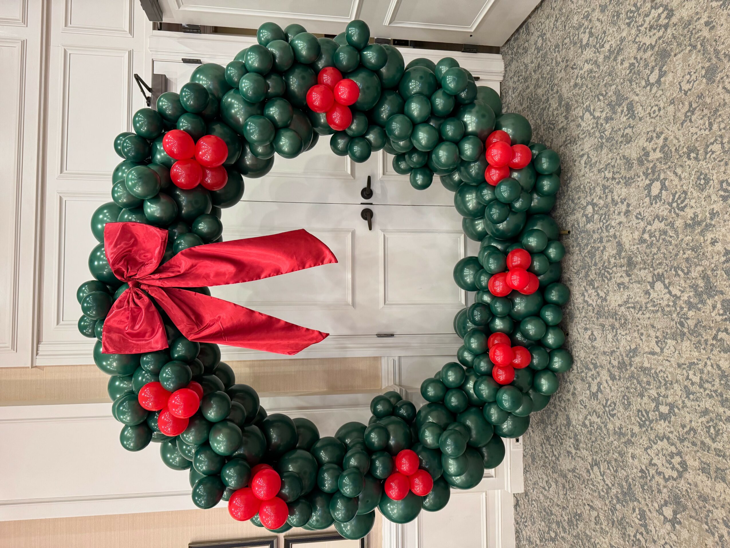 Christmas wreath made of green and red balloons with a red bow, hanging on a white cabinet door.
