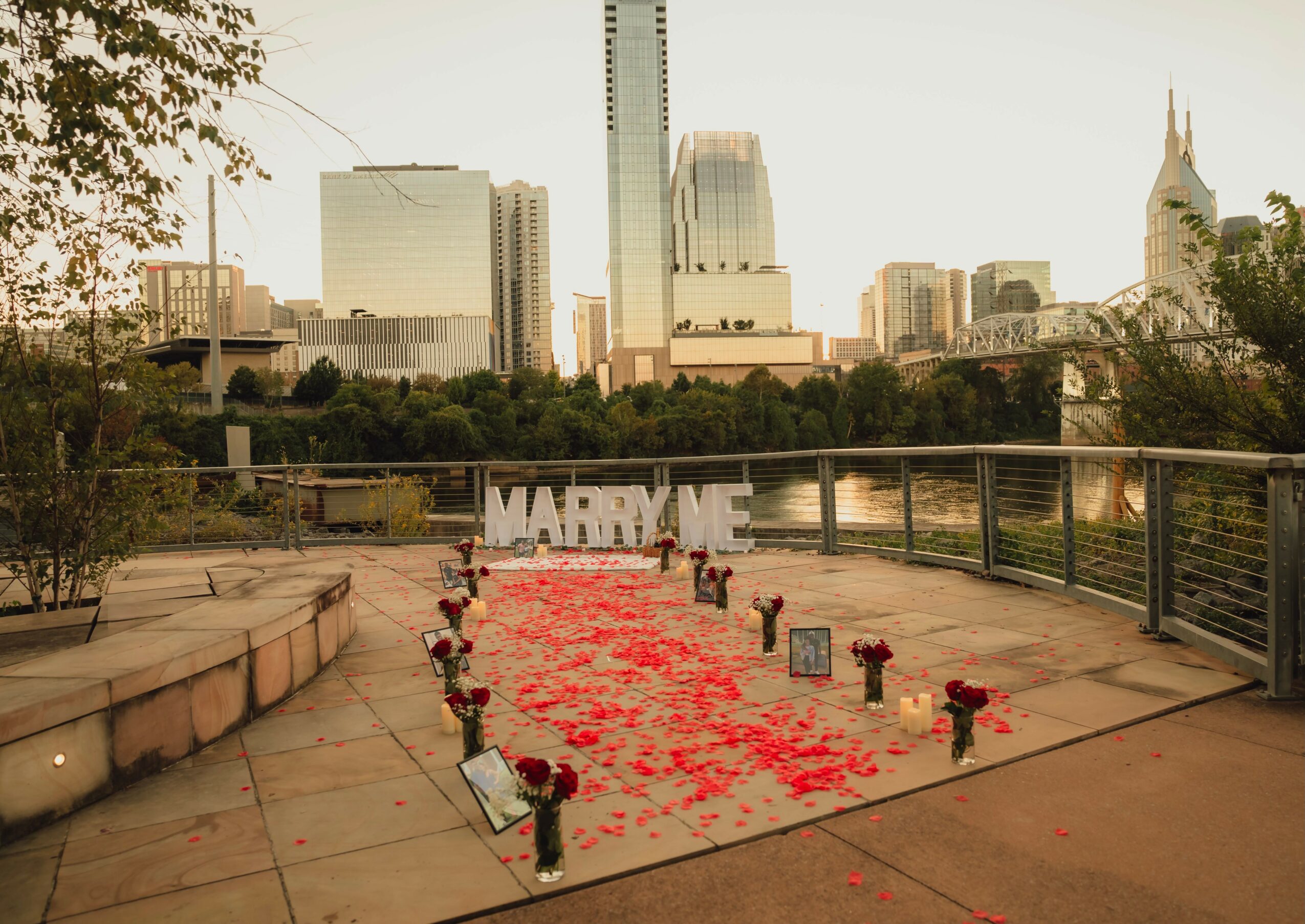 Memorial with red flower petals, small vases, and photos on a walkway overlooking a city skyline.