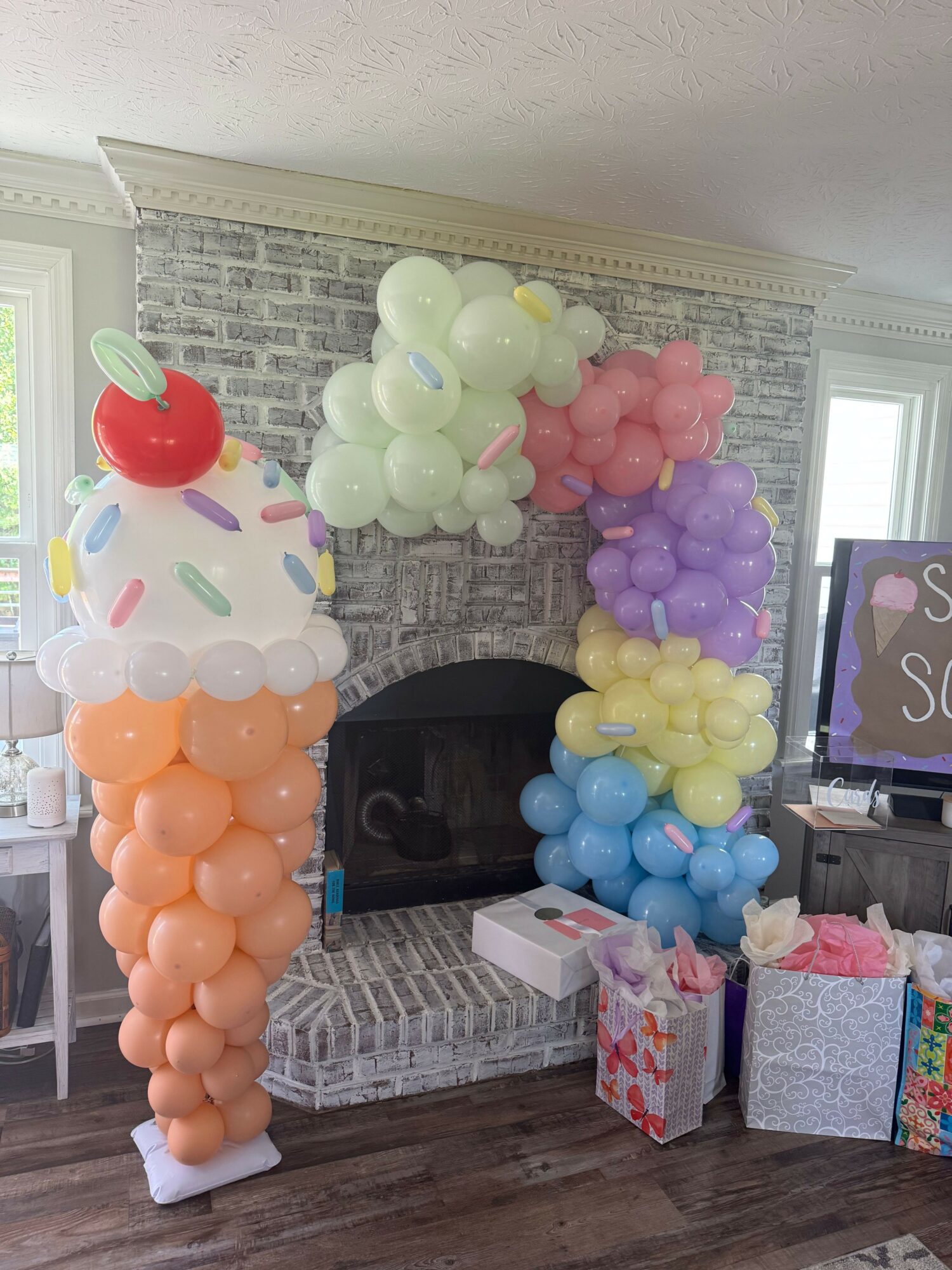 Balloon arrangement with various colors and a cake with balloons on top, in a room with a brick wall and fireplace.