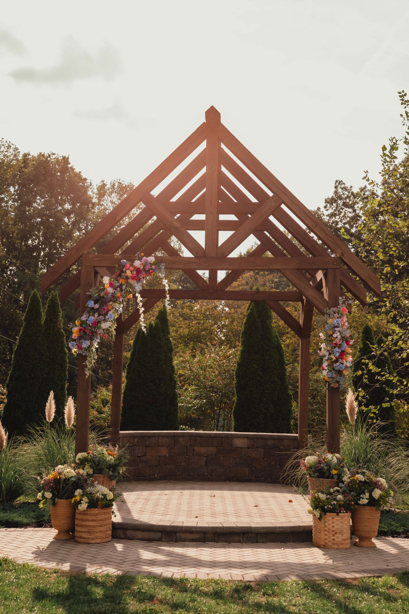 Wooden arch with floral decorations, set in a garden with trees and potted plants, under a cloudy sky.