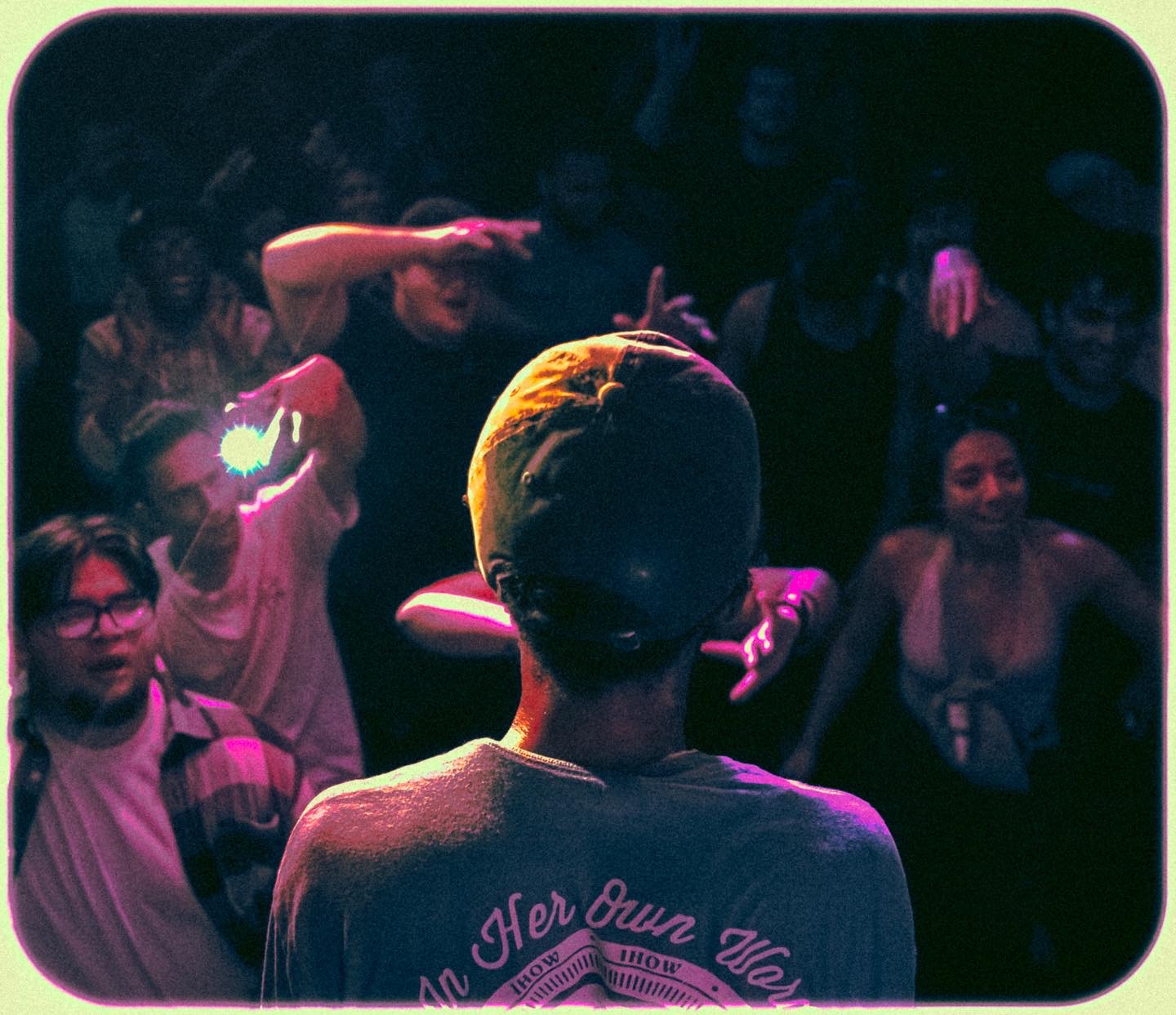 Person with a cap facing away, performing on stage with a crowd watching, some taking photos, in a dark setting.