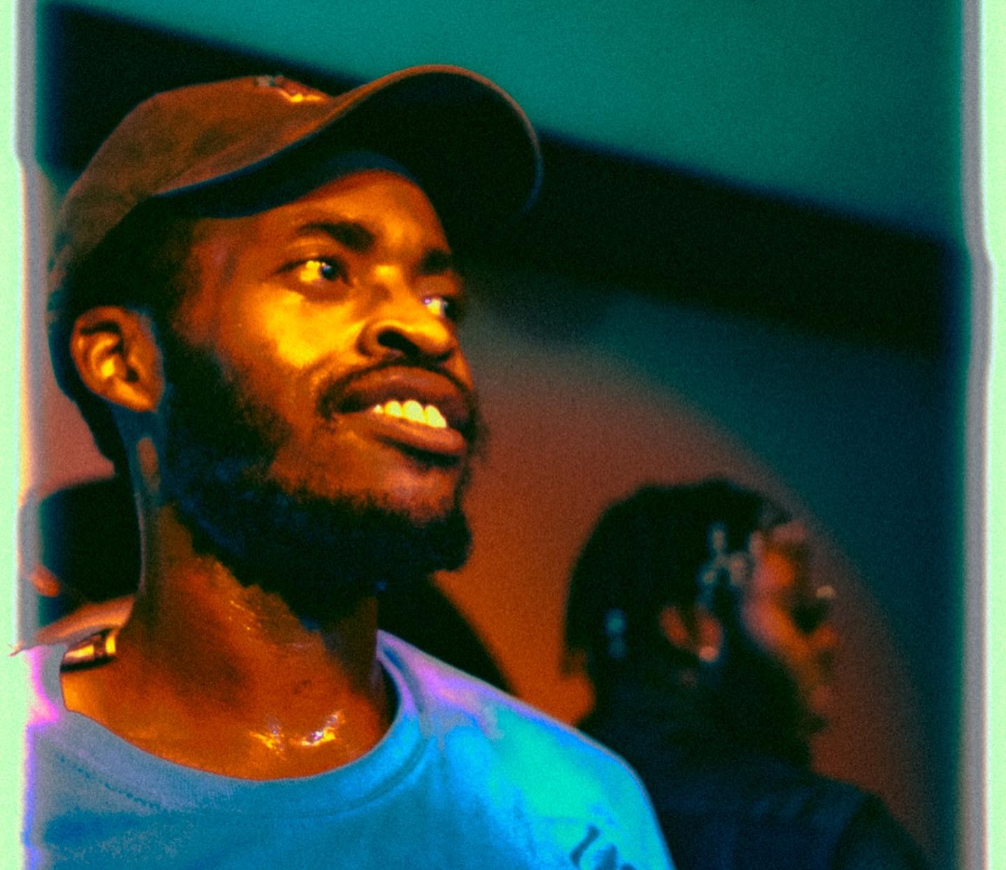 Young man with a beard and cap smiling, with a blurred person in the background, colorful lighting.