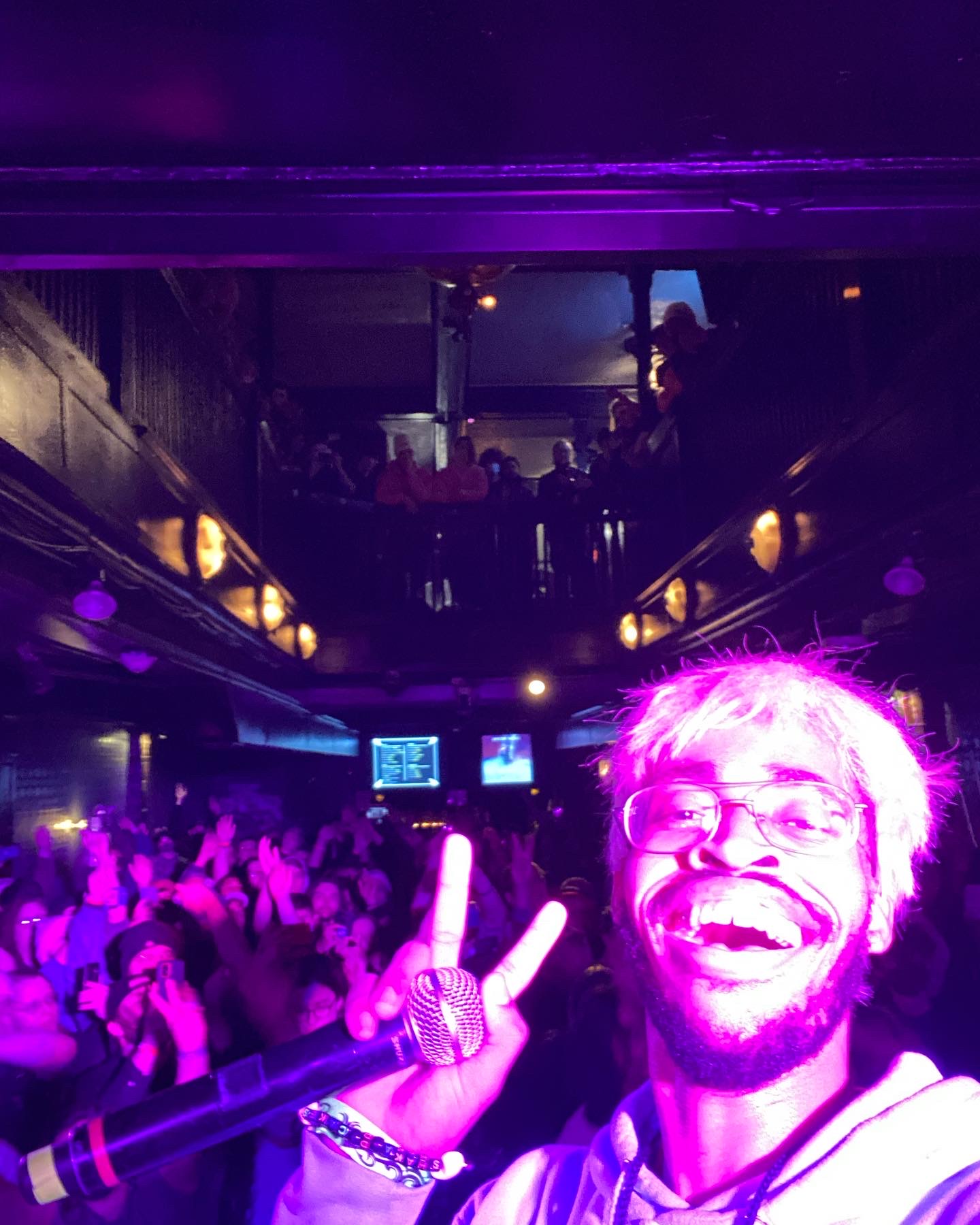 Person smiling and making a peace sign at a crowded indoor event with purple lighting.