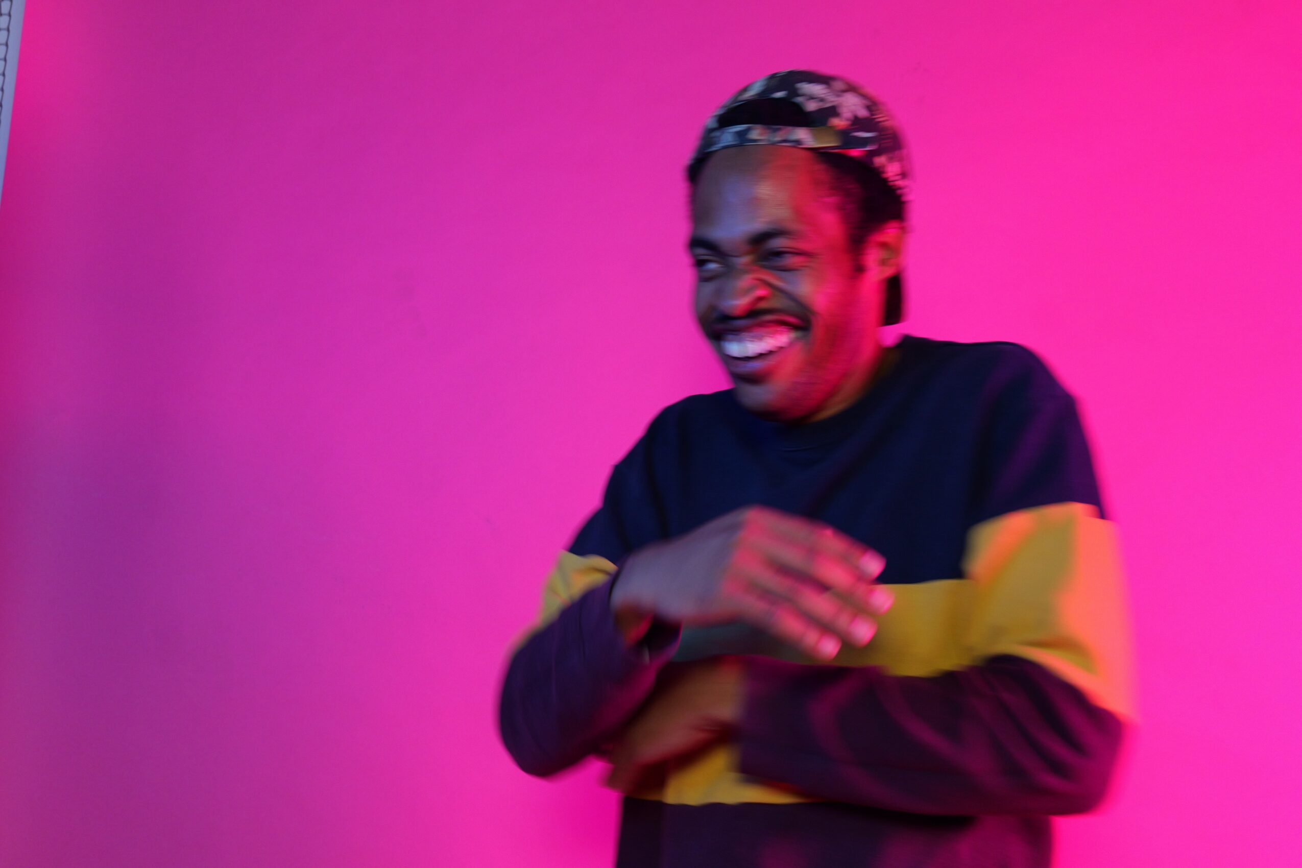 Person laughing with arms crossed, wearing a dark sweater with yellow and pink background.
