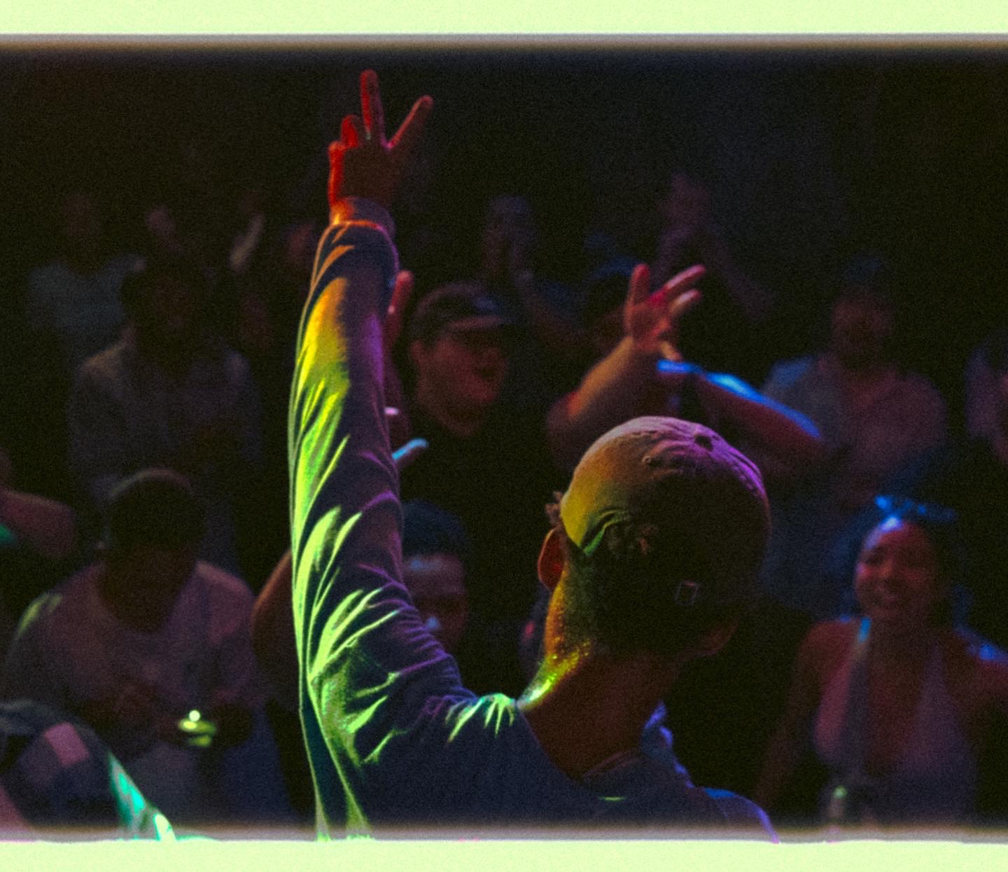 Person with short hair raising hand with peace sign in a dark, colorful environment with an audience.