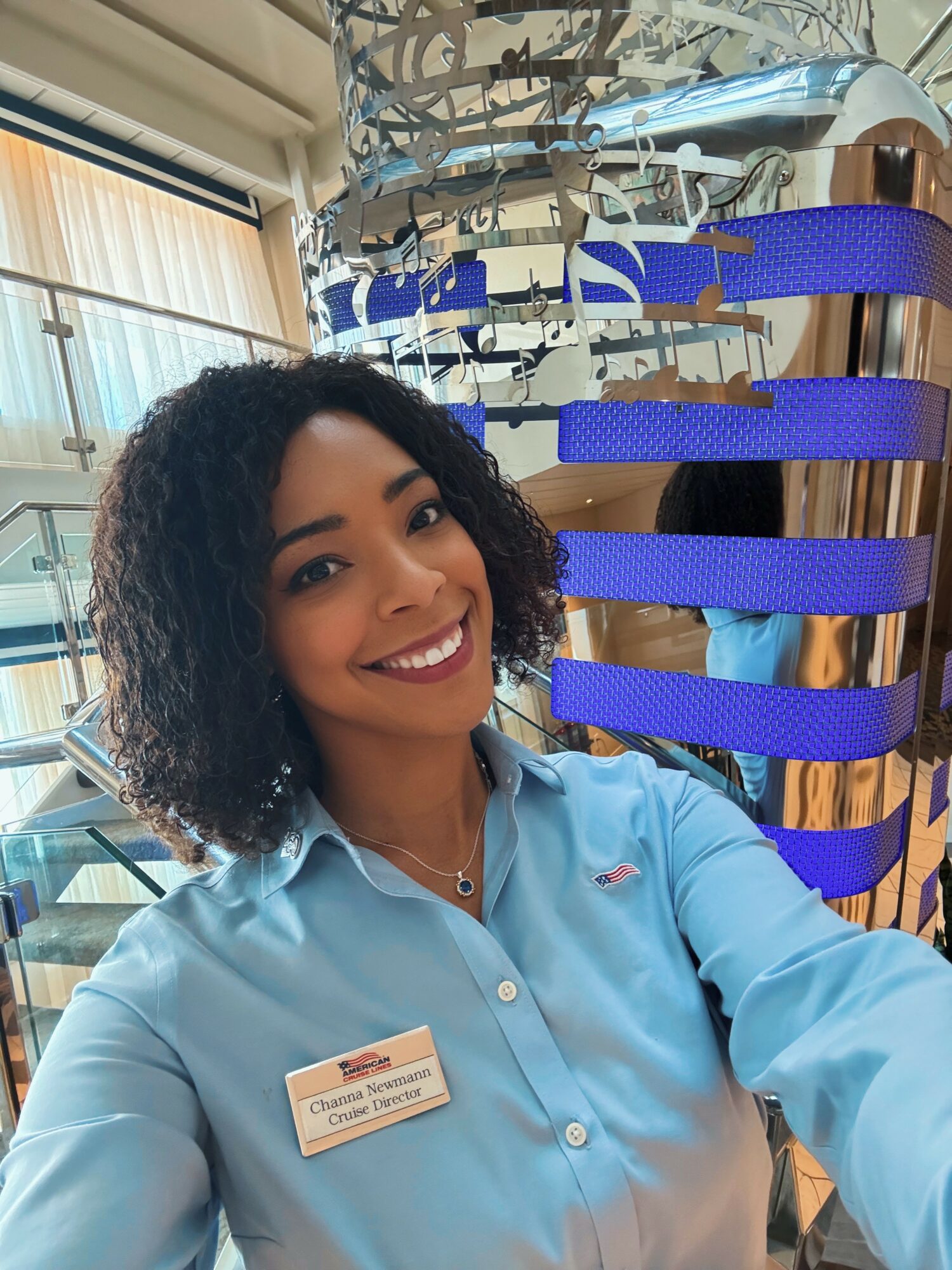 Smiling woman with curly hair in a light blue shirt, standing indoors near a display with blue straps and metallic objects.