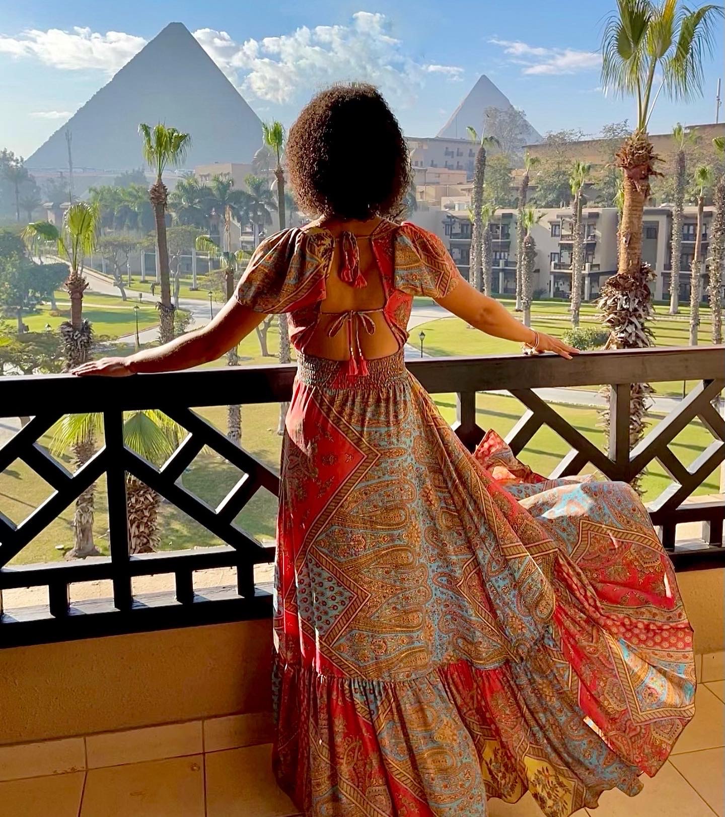Person with curly hair wearing a colorful dress, standing on a balcony overlooking pyramids and palm trees.